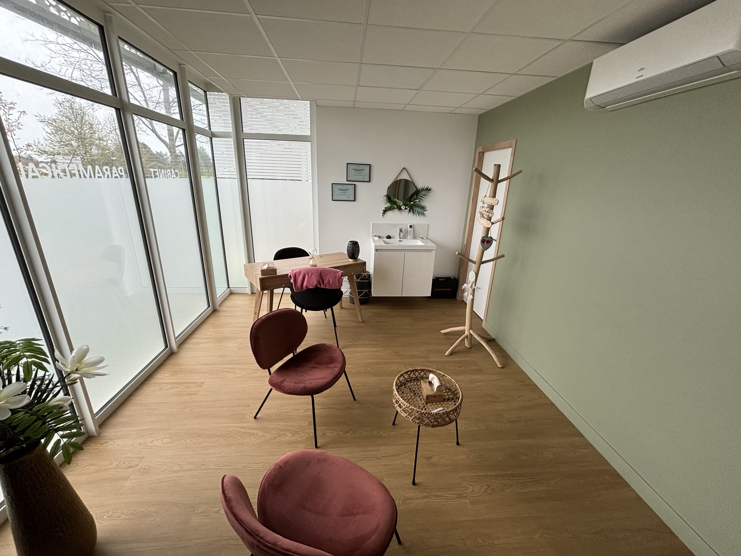Salle d'attente ou espace de consultation avec fauteuils en velours brodés, une table basse, un miroir et un lavabo avec un décor végétal, grande fenêtre en verre.