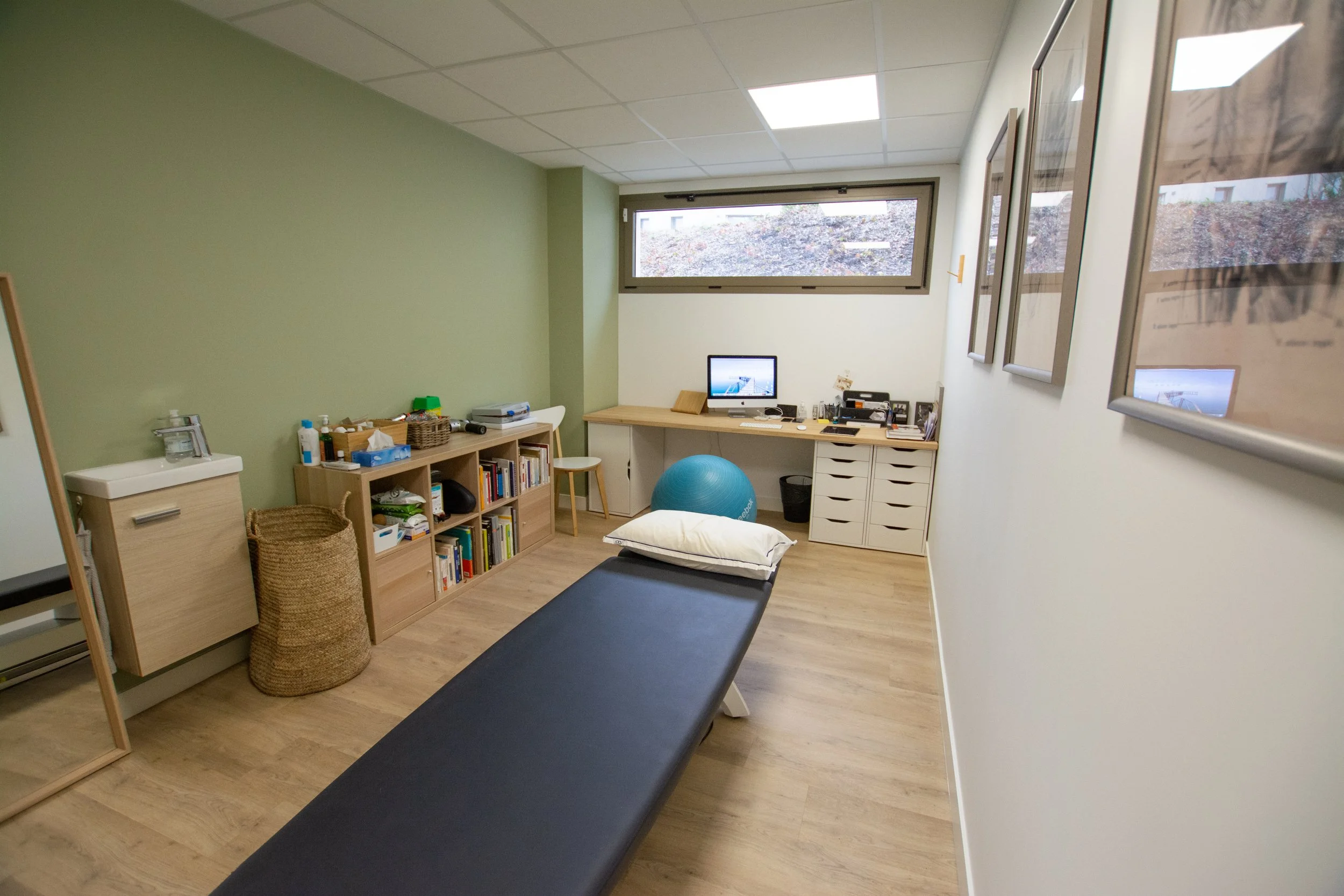 Salle de consultation avec un lit, un ballon de yoga bleu, une bibliothèque, un bureau avec un ordinateur, et des cadres suspendus au mur.