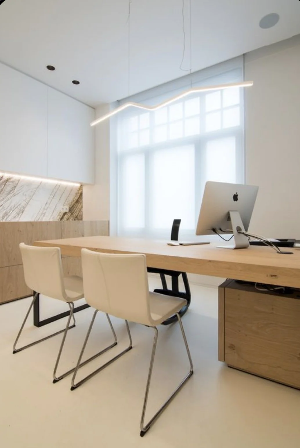 Bureau moderne avec un grand bureau en bois, deux chaises blanches, un ordinateur Apple sur le bureau, une grande fenêtre avec un rideau translucide, un mur en bois et un éclairage contemporain suspendu.