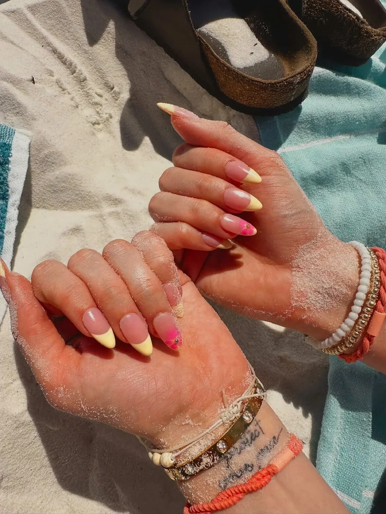 Close-up of well-manicured hands with French tip nails and pink accent, resting on sandy beach with beach shoes in the background, wearing colorful bracelets and jewelry.