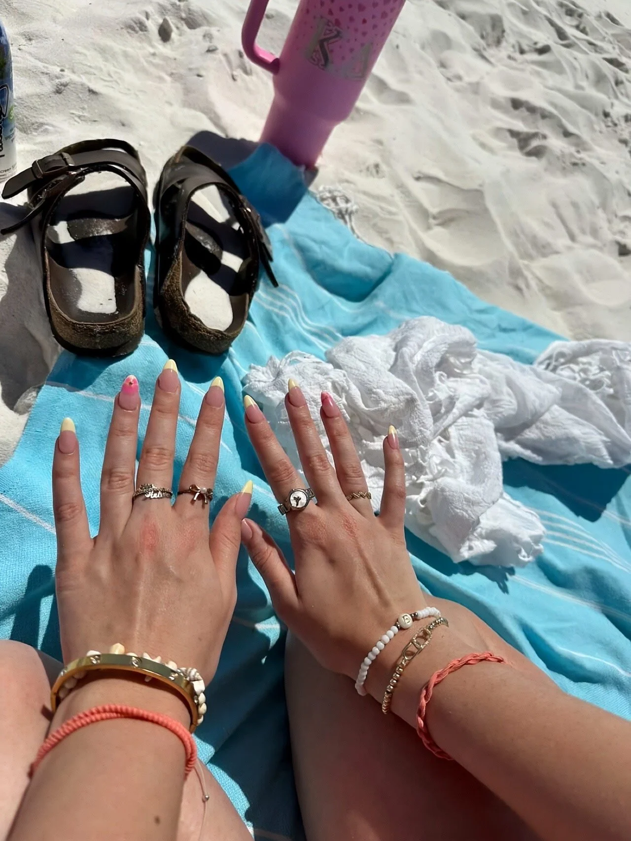 Two hands with decorated fingernails and jewelry resting on a blue towel on white sand, with sandals, a pink water bottle, and white cloth nearby.