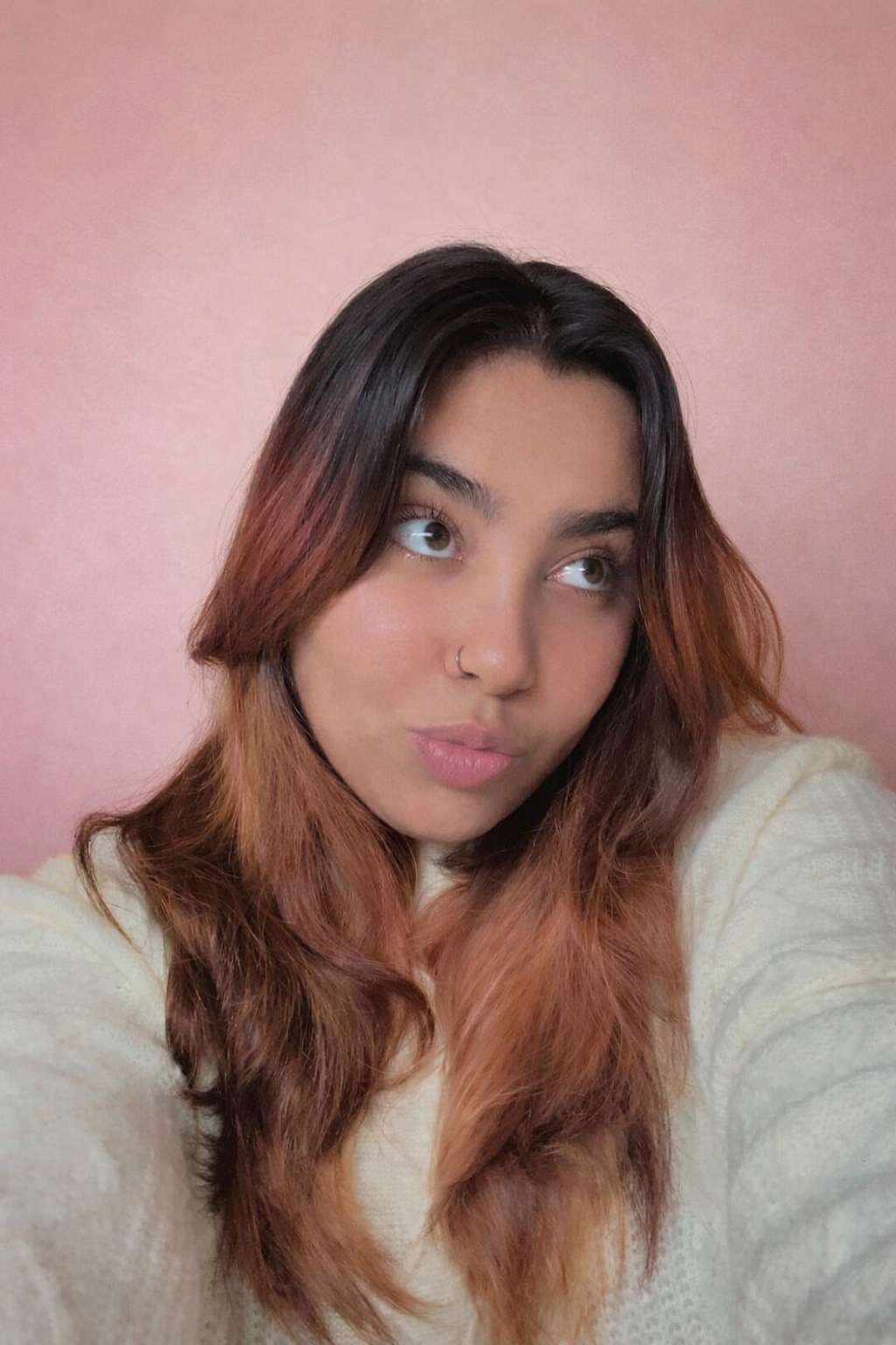 A young woman with brown to reddish hair and light skin, wearing a cream-colored sweater, looking upwards with a pink background.