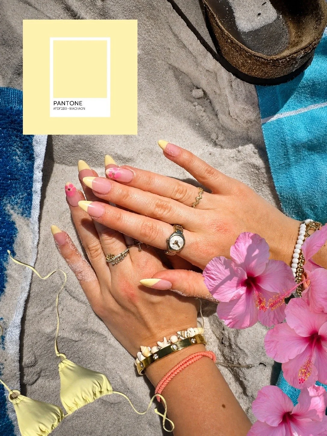 Two hands on sandy beach with pink hibiscus flowers, colorful bracelets, rings, and yellow and pink nail polish, next to a blue towel and a drink in a coconut.