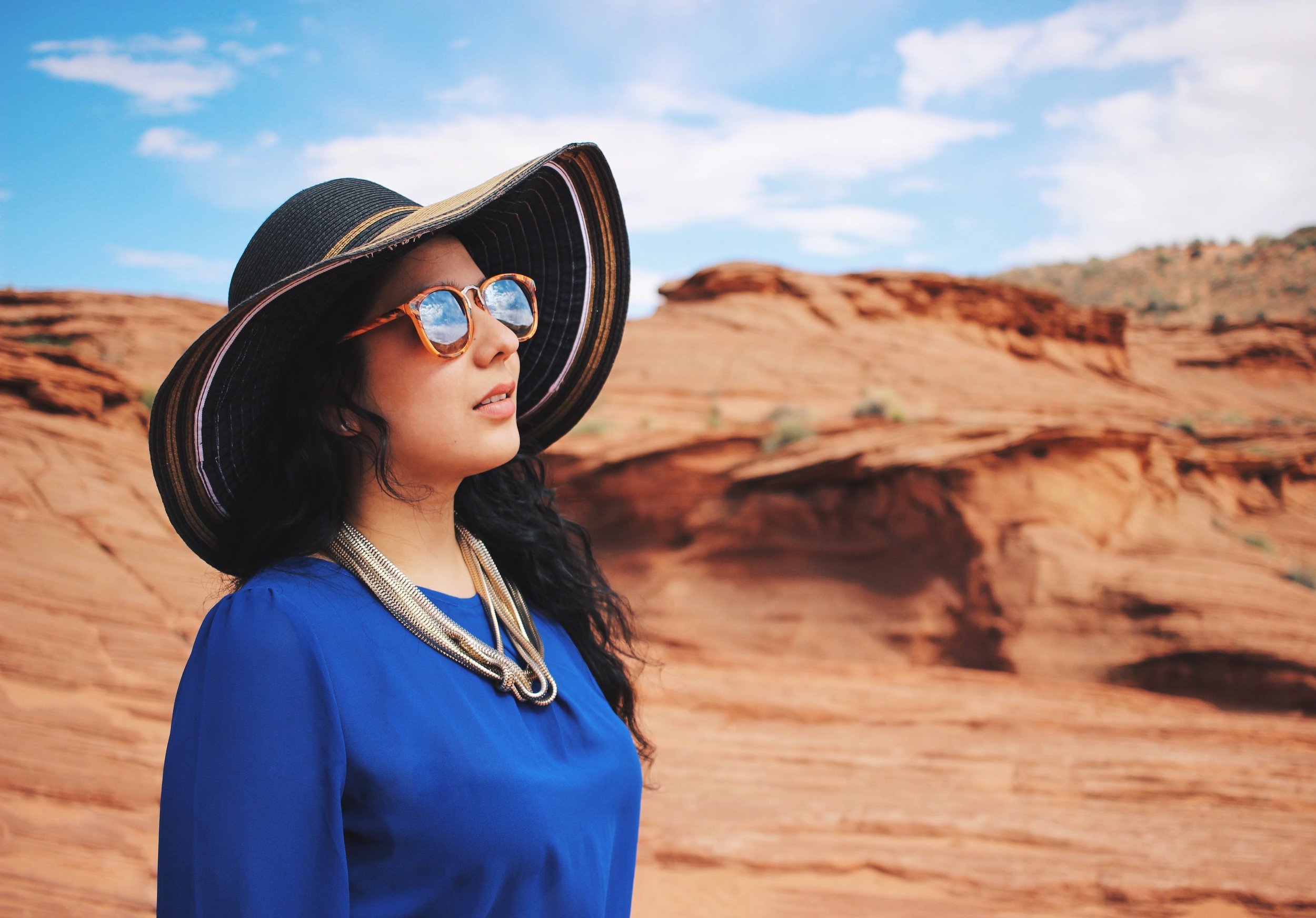 Woman wearing a large sun hat, sunglasses, and a blue top in a desert landscape with red rocks and a blue sky.