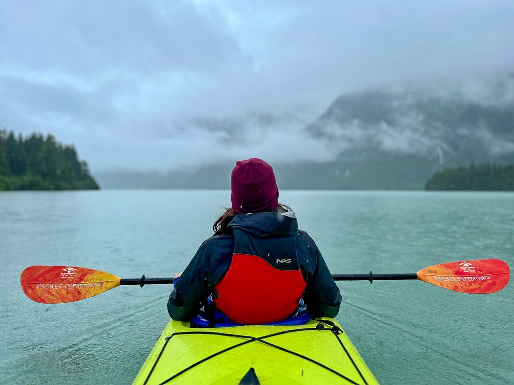 Person kayaking on a large body of water with mountainous and forested landscape in the background, overcast sky, wearing a maroon beanie, dark jacket, and red life vest.