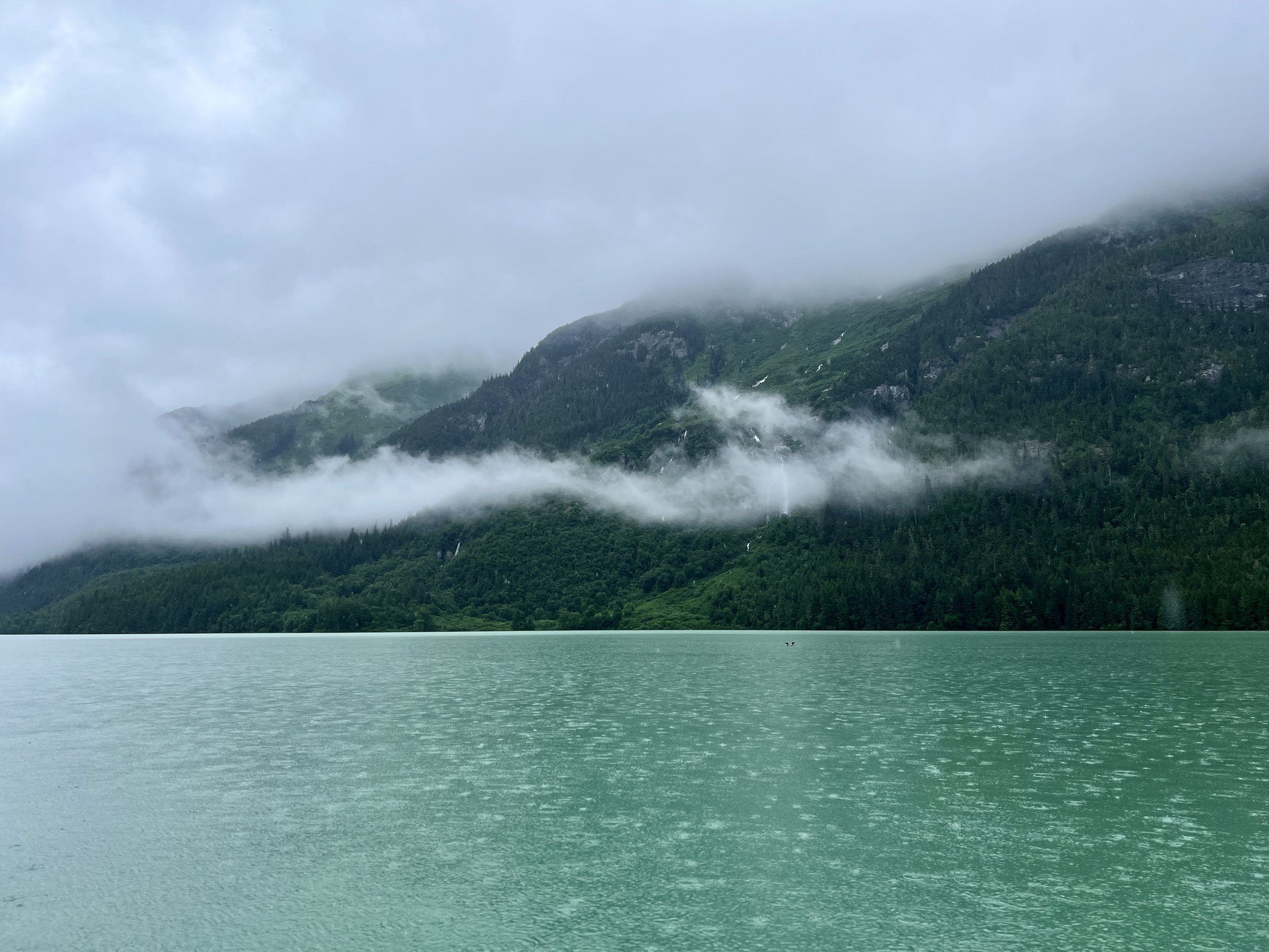 A calm teal lake with misty green mountains and dense forest in the background, shrouded in low clouds