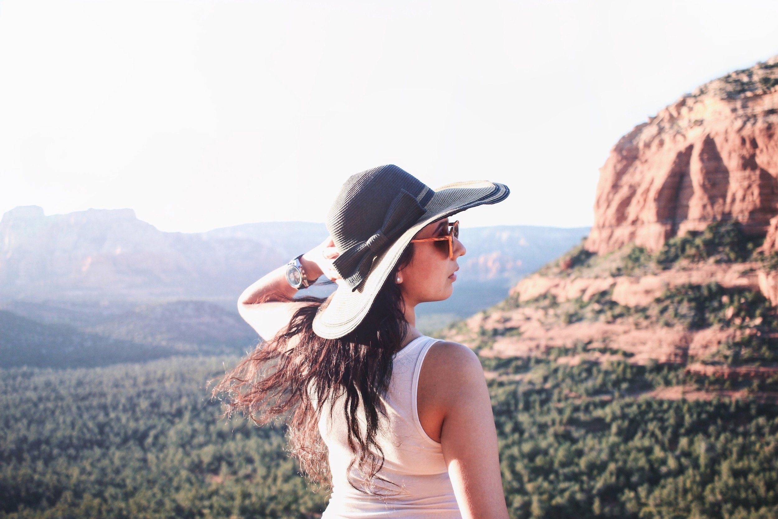 A woman wearing sunglasses, a wide-brimmed hat, and a sleeveless top looks into the distance at a scenic mountainous landscape with red rock formations and green forested areas.