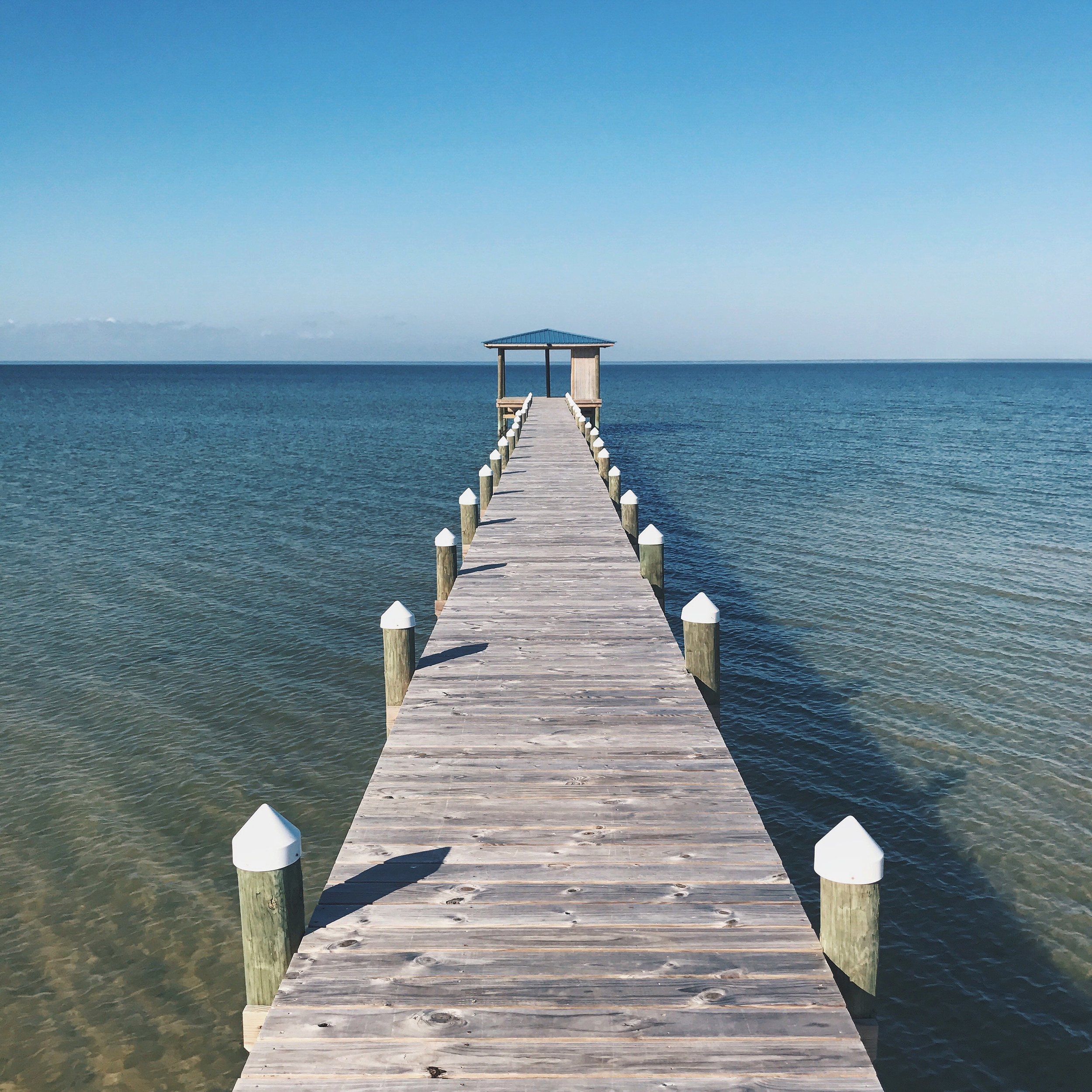 Wooden pier extending into the ocean with a small shelter at the end, under a clear blue sky.
