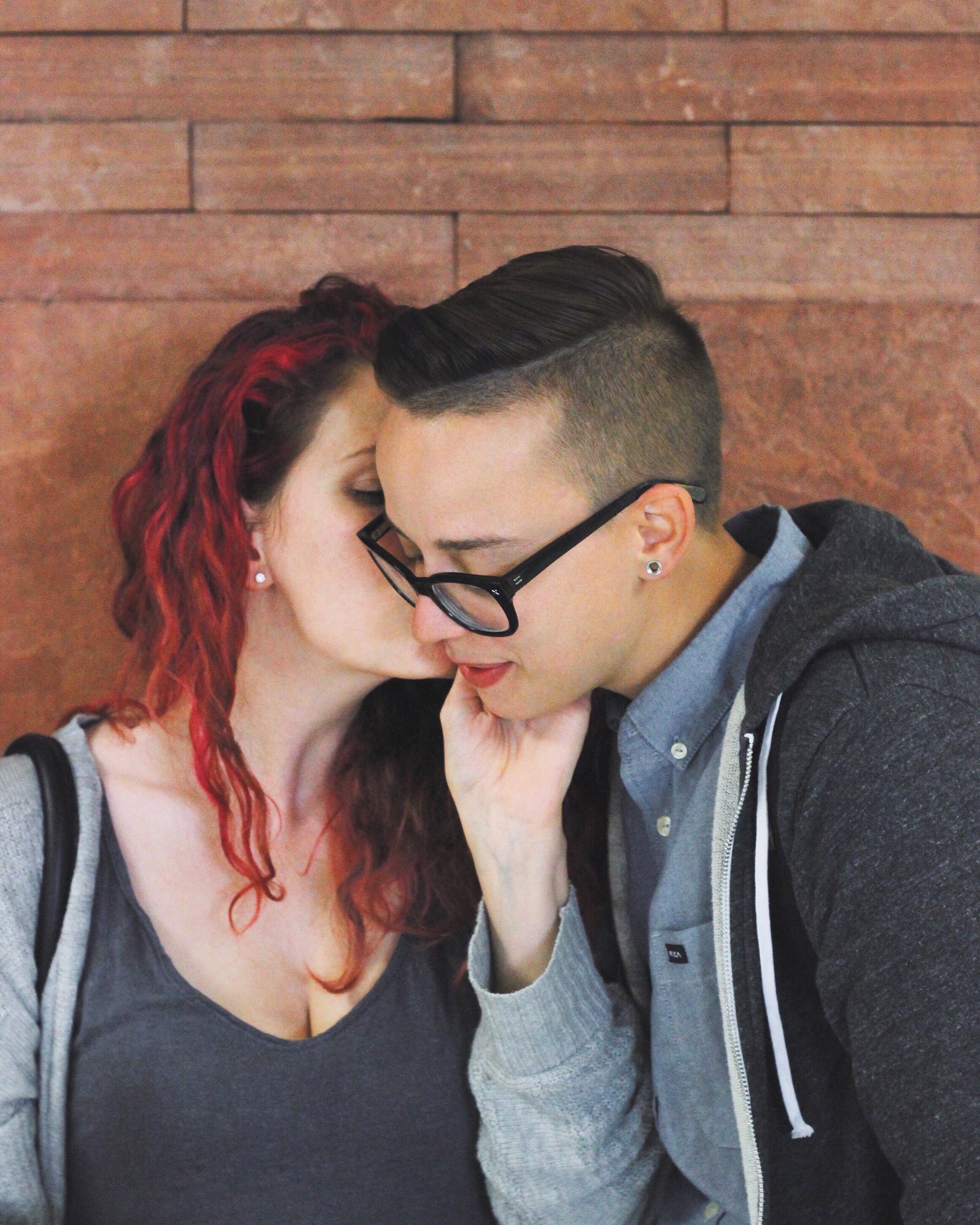 Two women sharing a close, intimate moment with their foreheads touching, one with curly red hair and the other with short dark hair and glasses, against a wooden wall.