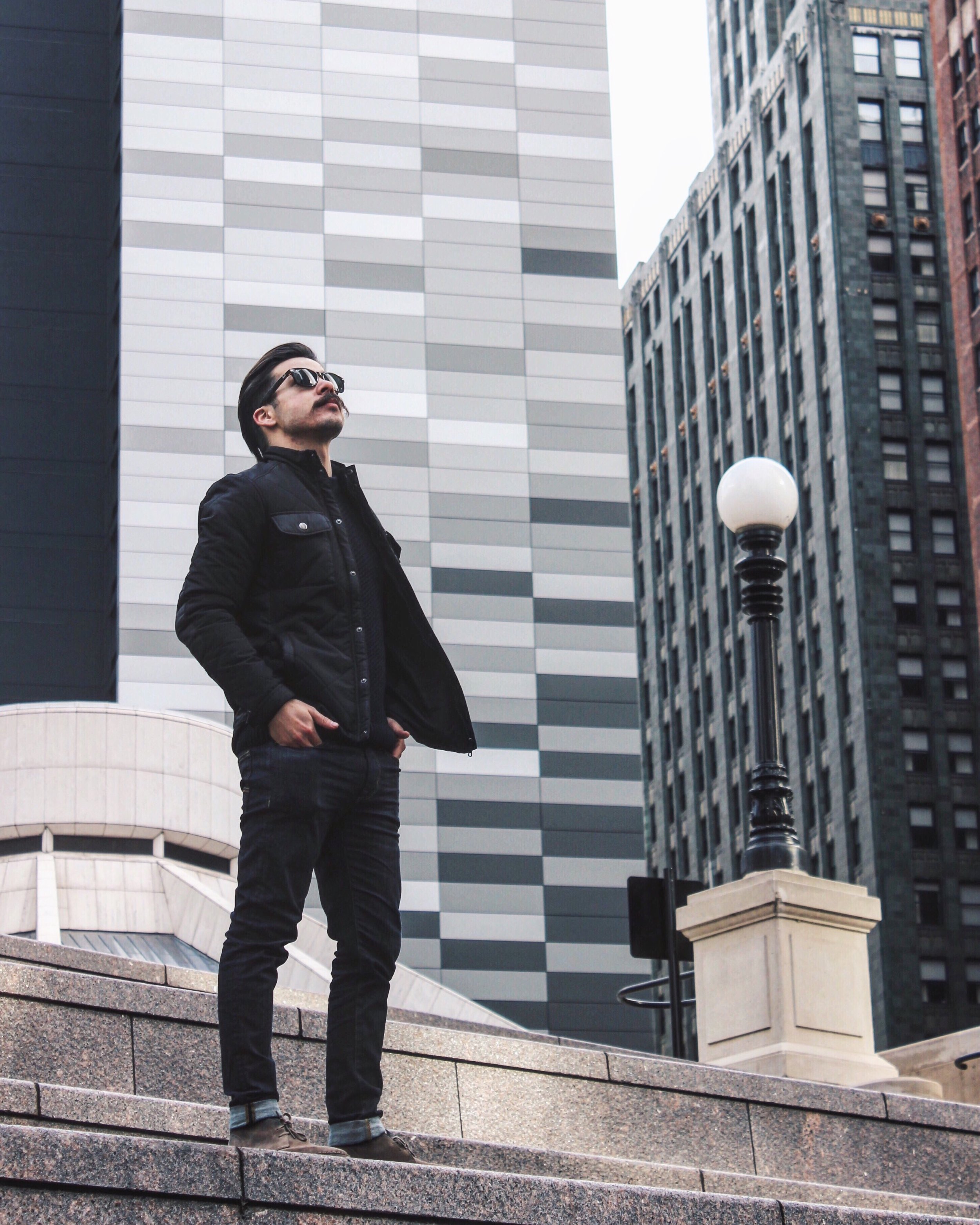 A man in black clothing and sunglasses standing on steps in an urban cityscape with tall modern buildings and a street lamp.
