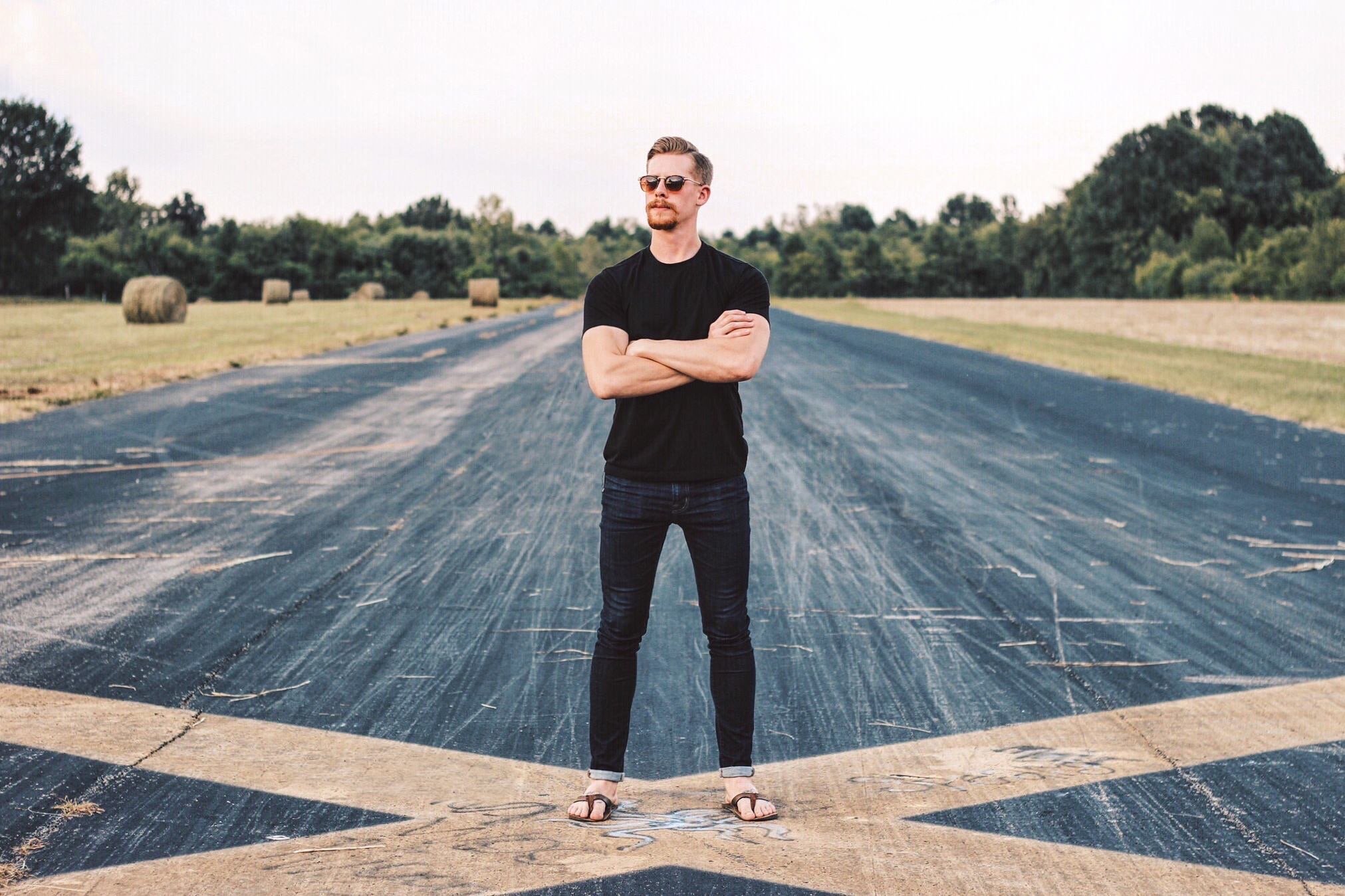 A man standing with arms crossed on a blacktop road in a rural area with hay bales and trees in the background.