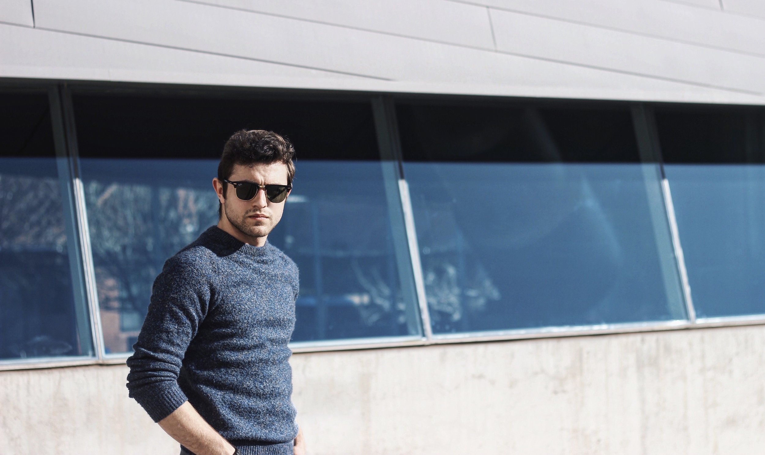 Young man with dark hair, wearing sunglasses and a blue sweater, standing outside near a modern building with large glass windows.