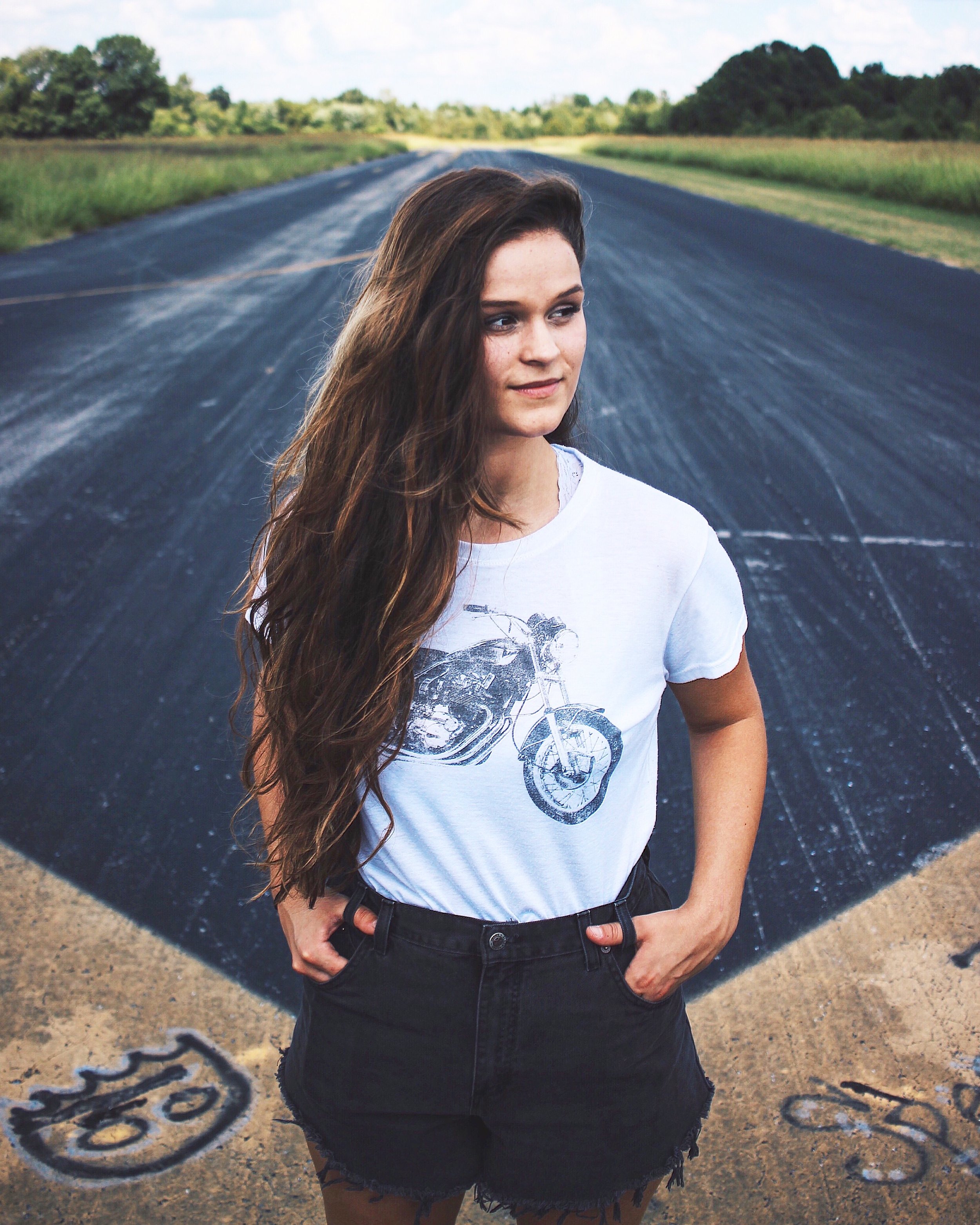 A young woman with long brown hair standing on a dirt road with a blacktop section behind her, surrounded by green fields and trees, under a partly cloudy sky.