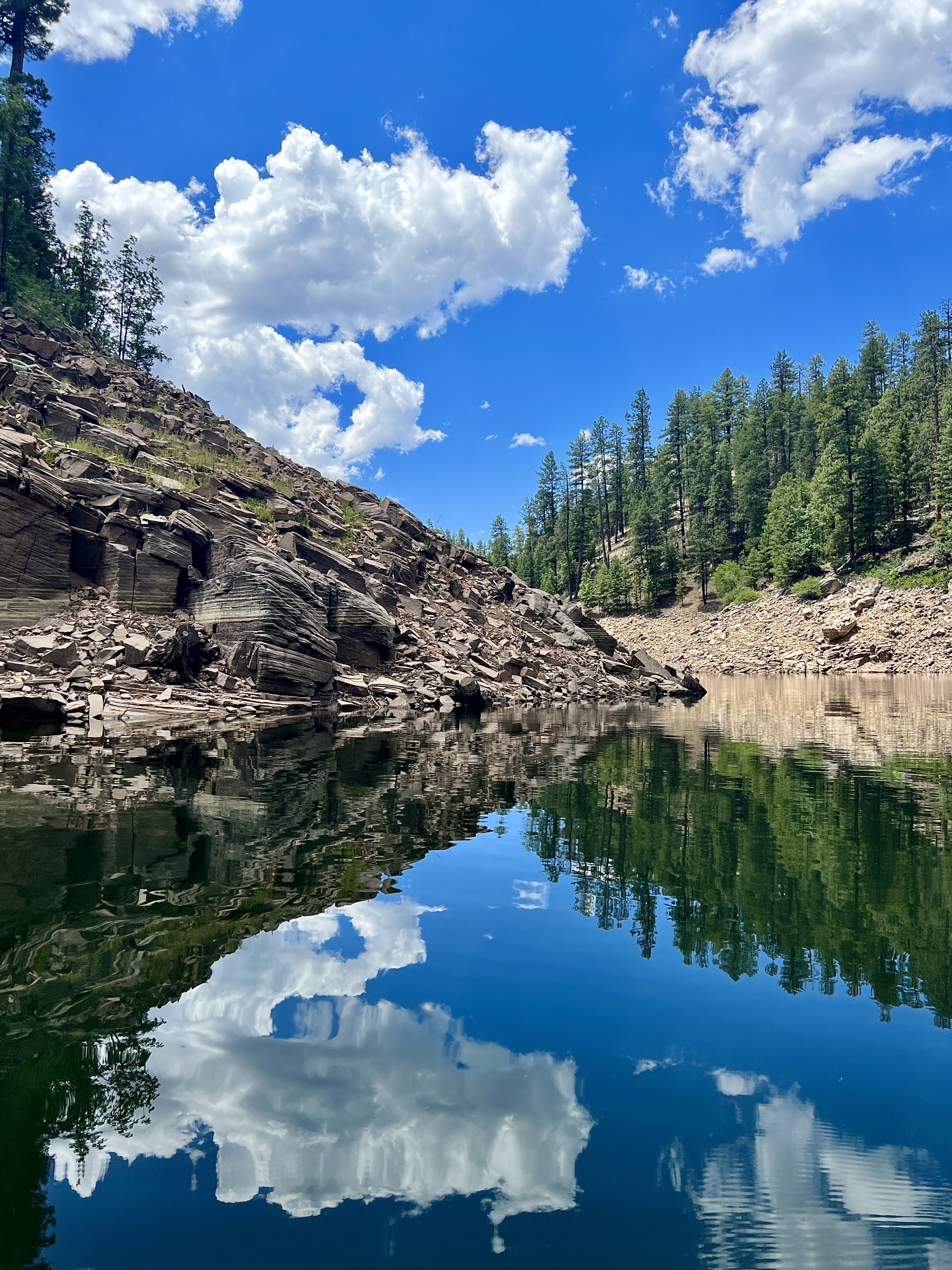 A serene mountain landscape with a clear blue sky and white clouds reflected in a calm river, surrounded by green pine trees and rocky slopes.