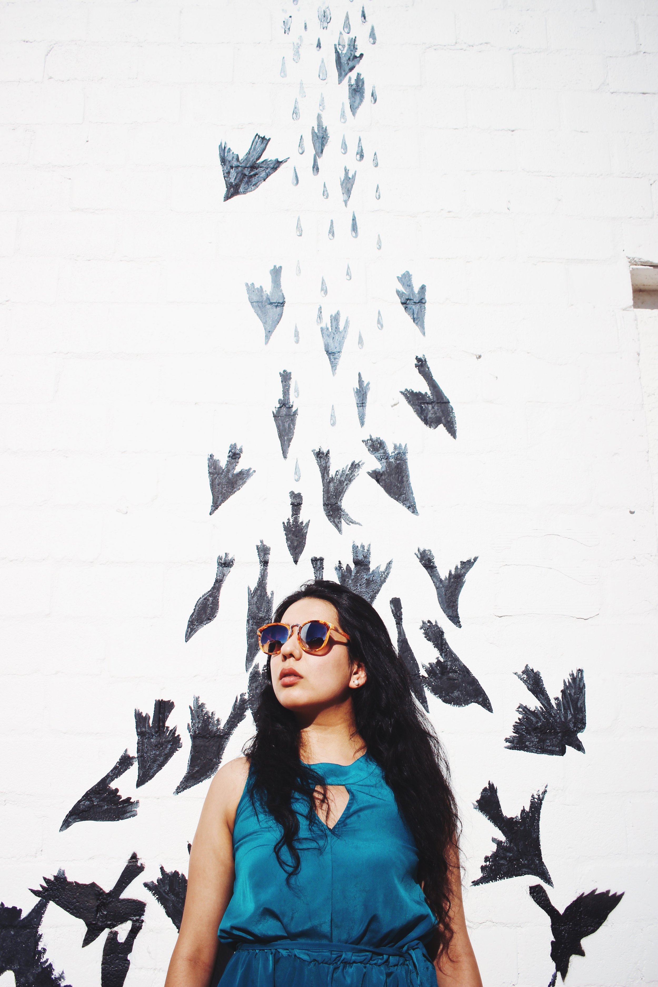 A woman with dark hair, wearing sunglasses and a blue dress, standing in front of a white brick wall with black bird illustrations flying upward.