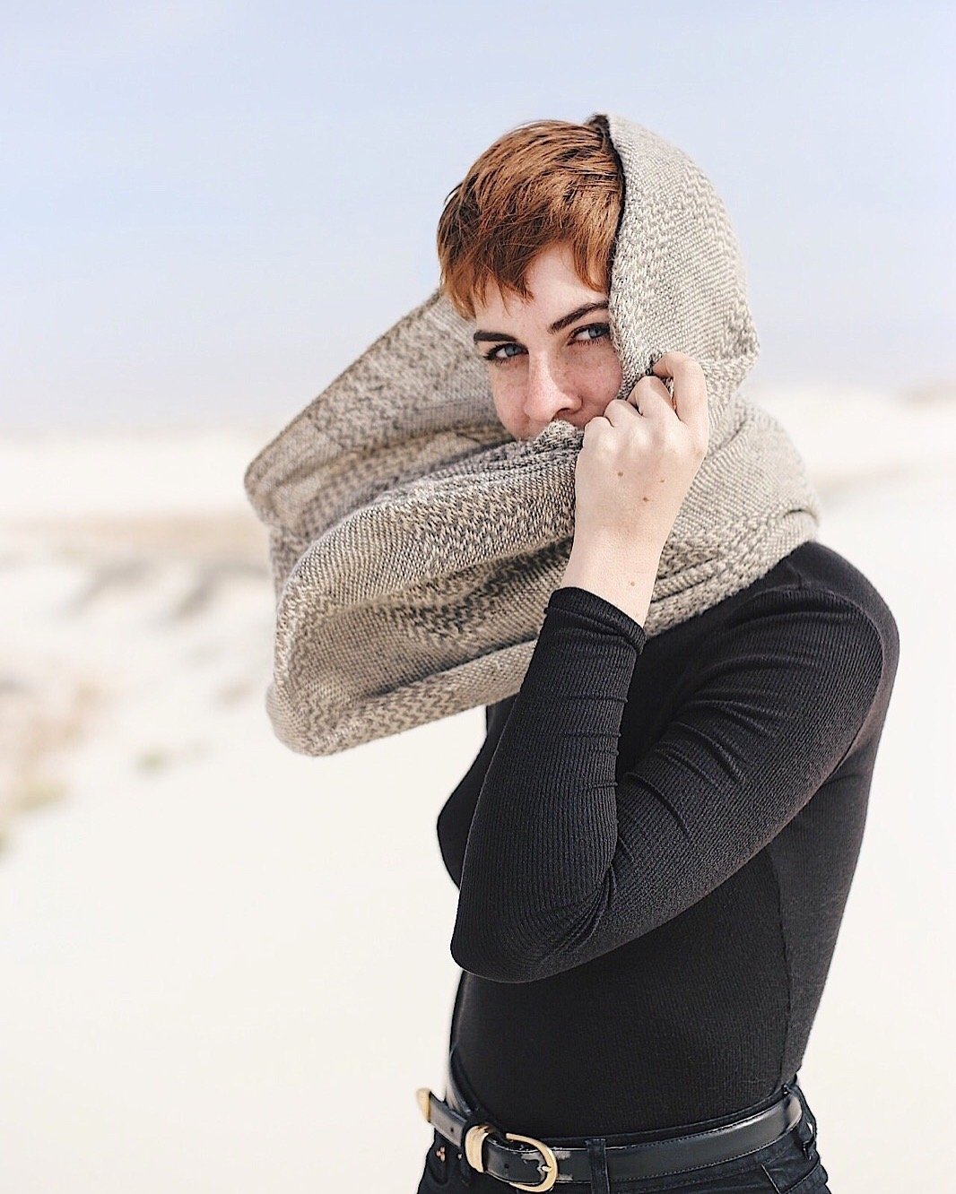A woman with short red hair and light skin standing outdoors in a snowy landscape, wrapped in a large beige scarf, wearing a black long-sleeve top and black pants, holding part of the scarf close to her face, with a slight smile.