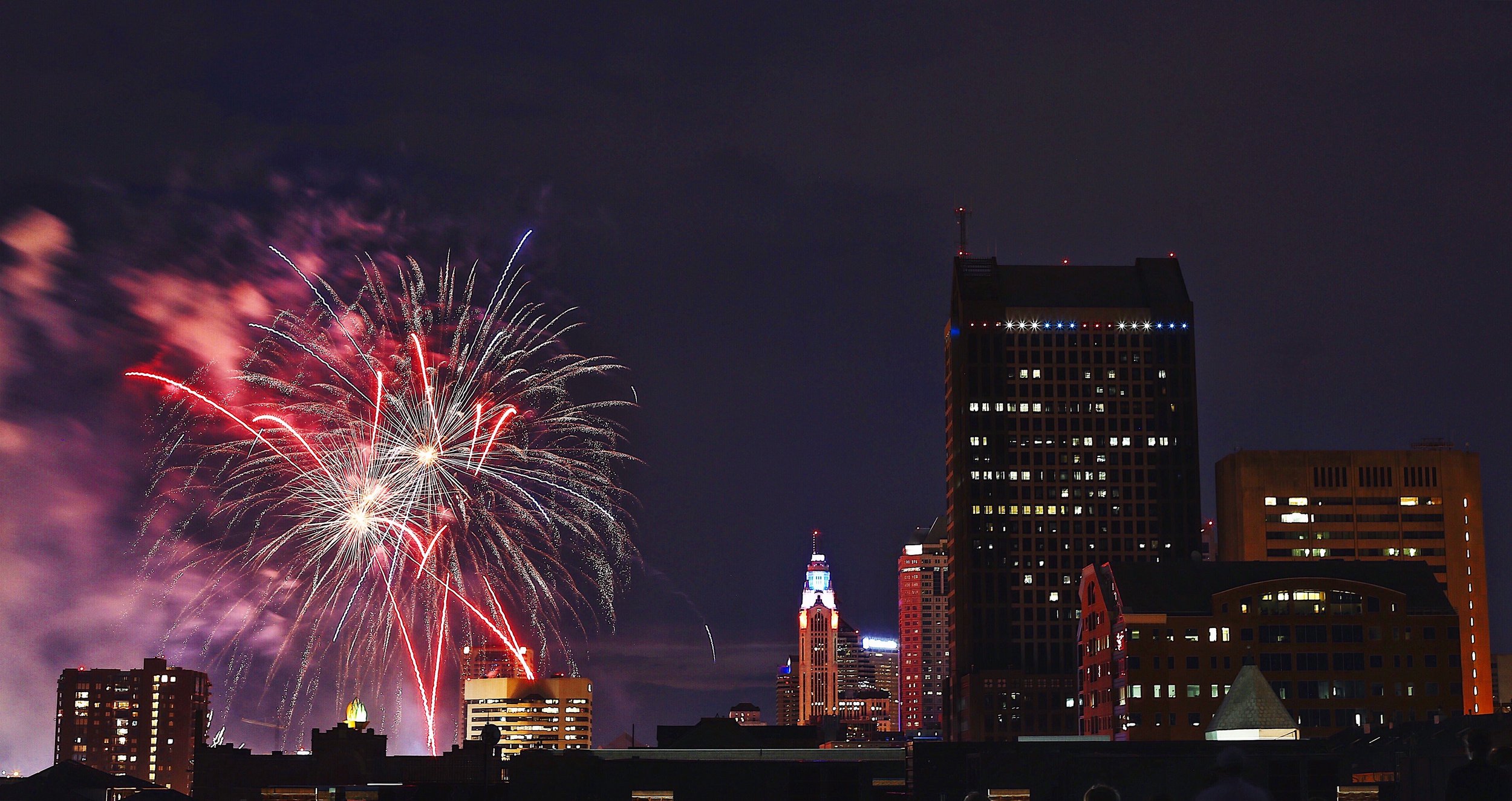 Fireworks exploding over a city skyline at night with illuminated buildings and a dark sky.
