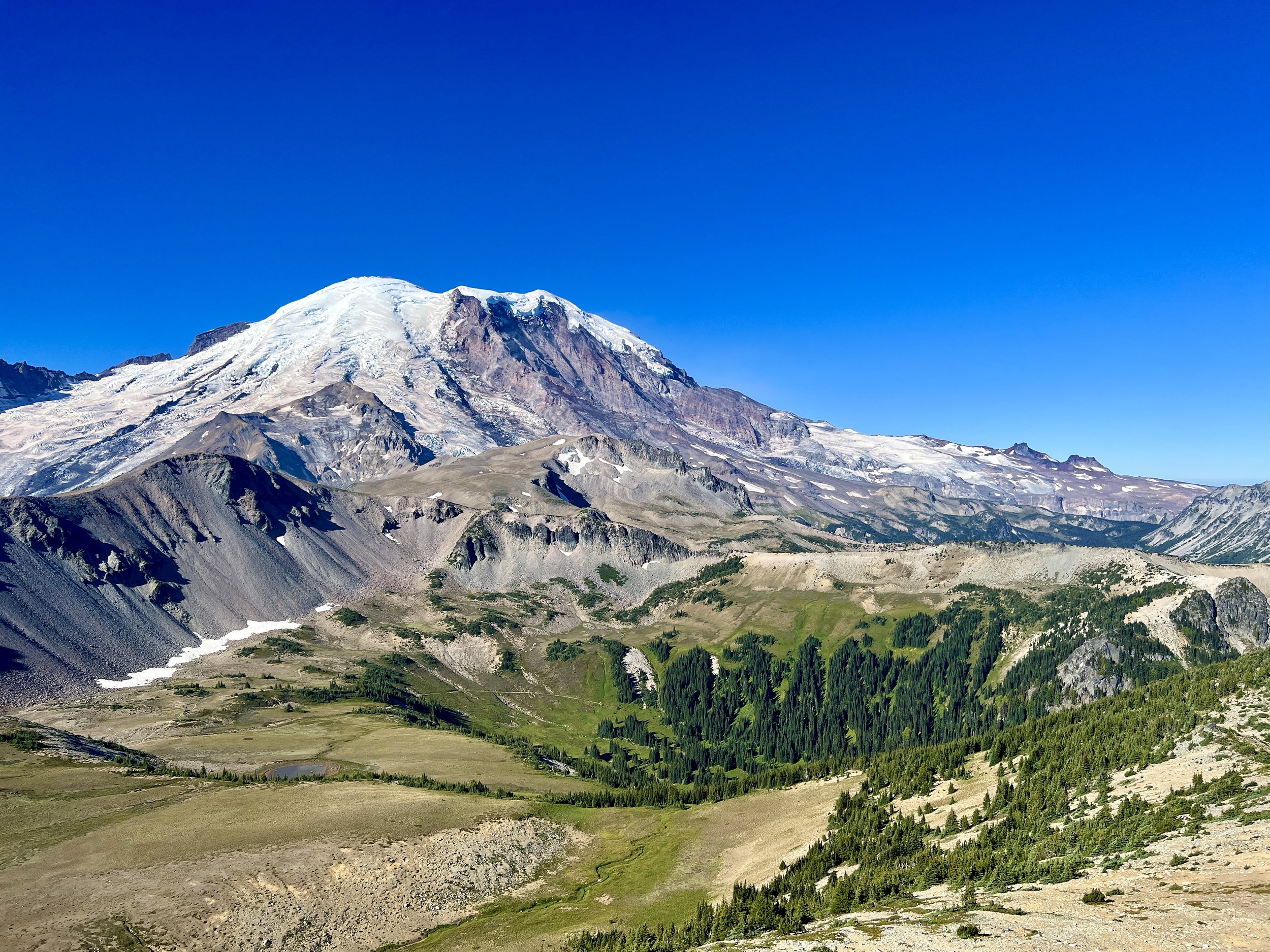 Snow-capped mountain with rocky slopes and a green valley below, under a clear blue sky.