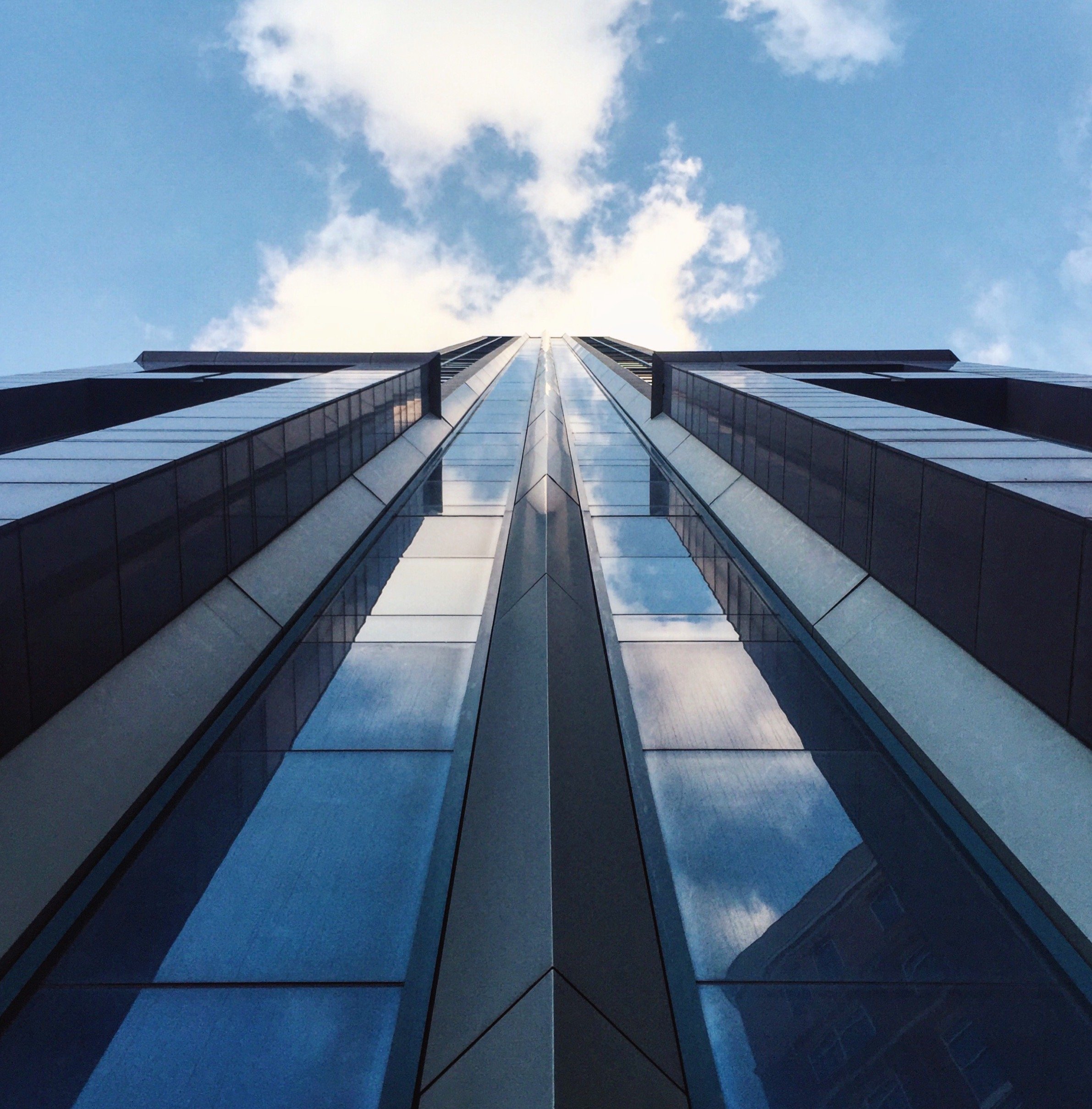 Looking up at a tall glass skyscraper with reflective windows against a blue sky with scattered clouds.