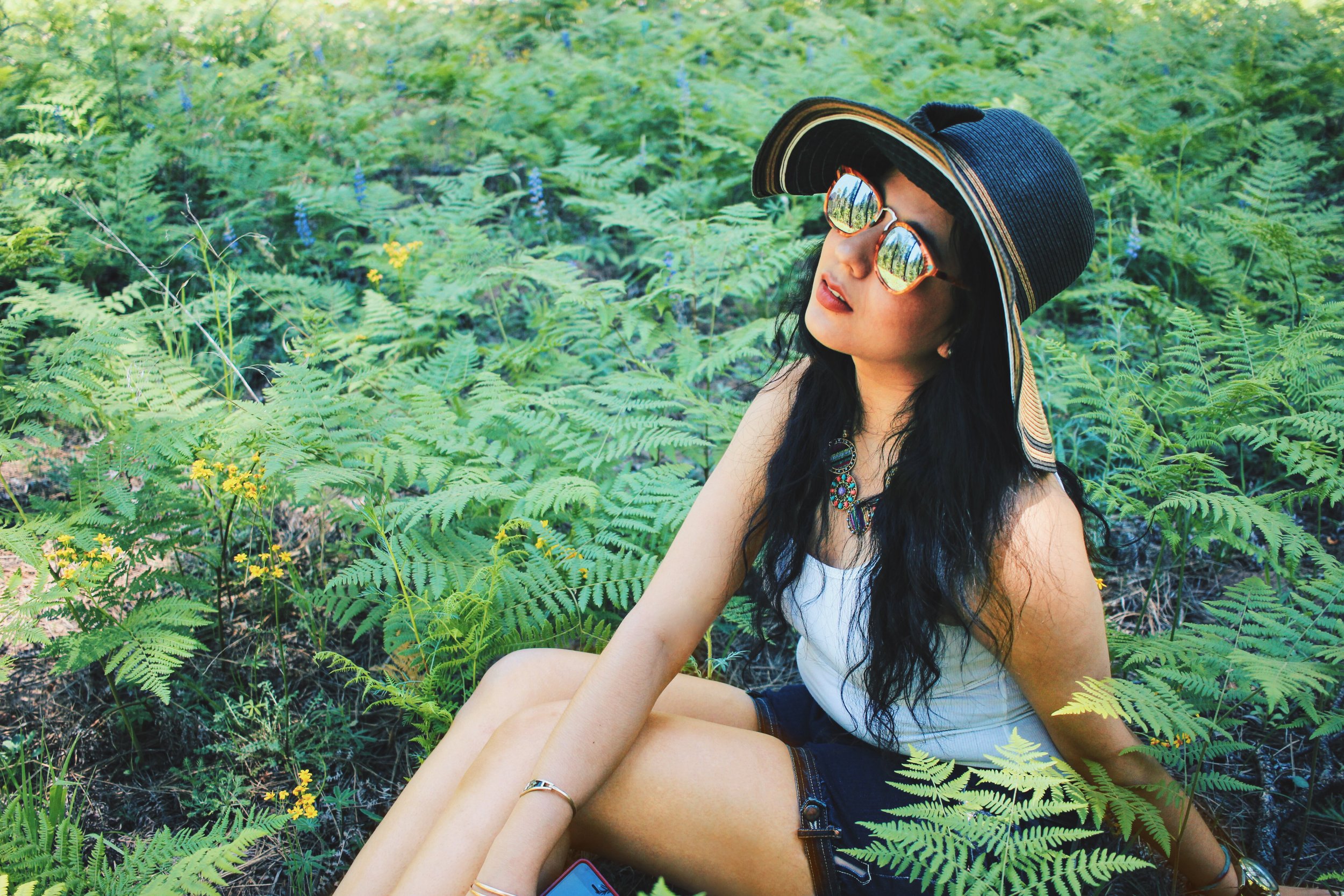A woman with long dark hair sitting among green ferns, wearing a wide-brimmed black sun hat with a striped band, round sunglasses reflecting the foliage, a colorful necklace, a white sleeveless top, and dark shorts.