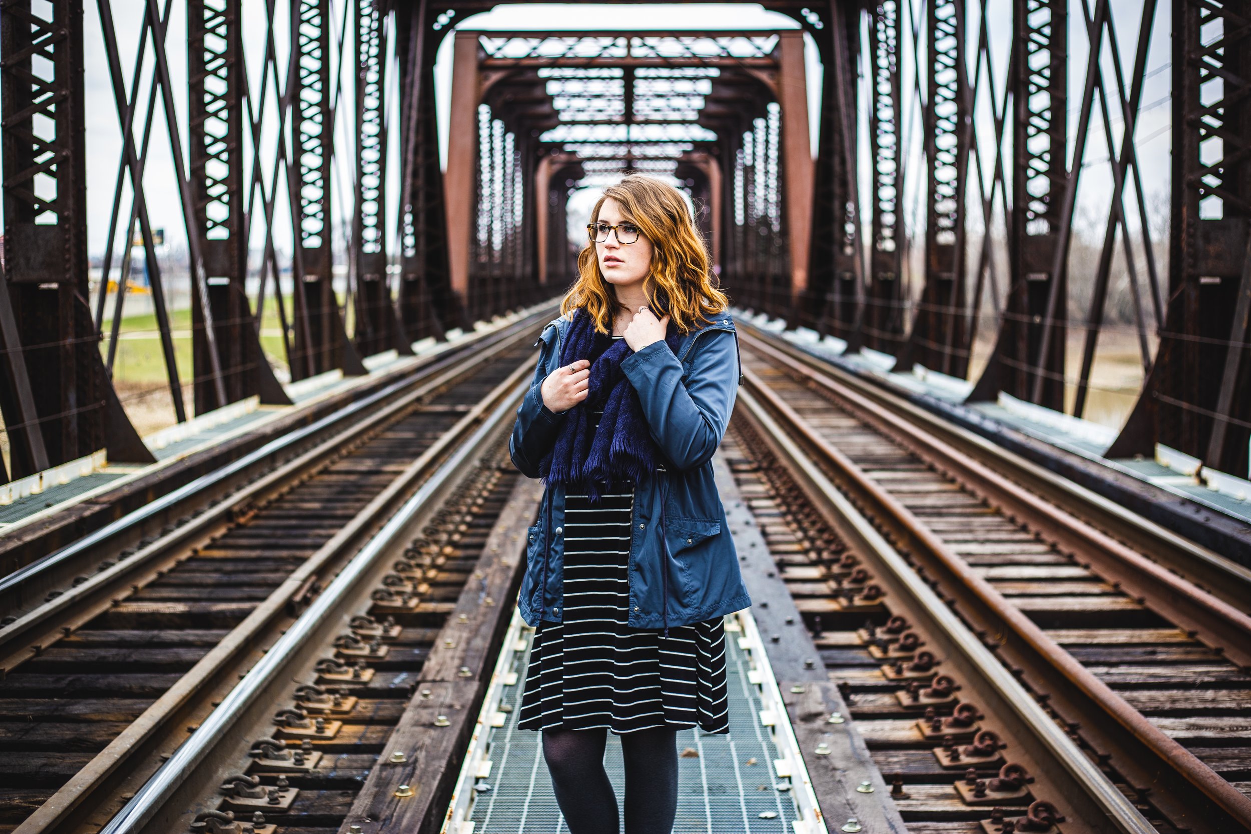 A young woman with red hair, glasses, and a blue jacket standing on a train bridge with railroad tracks, holding her jacket near her neck, looking to the side.