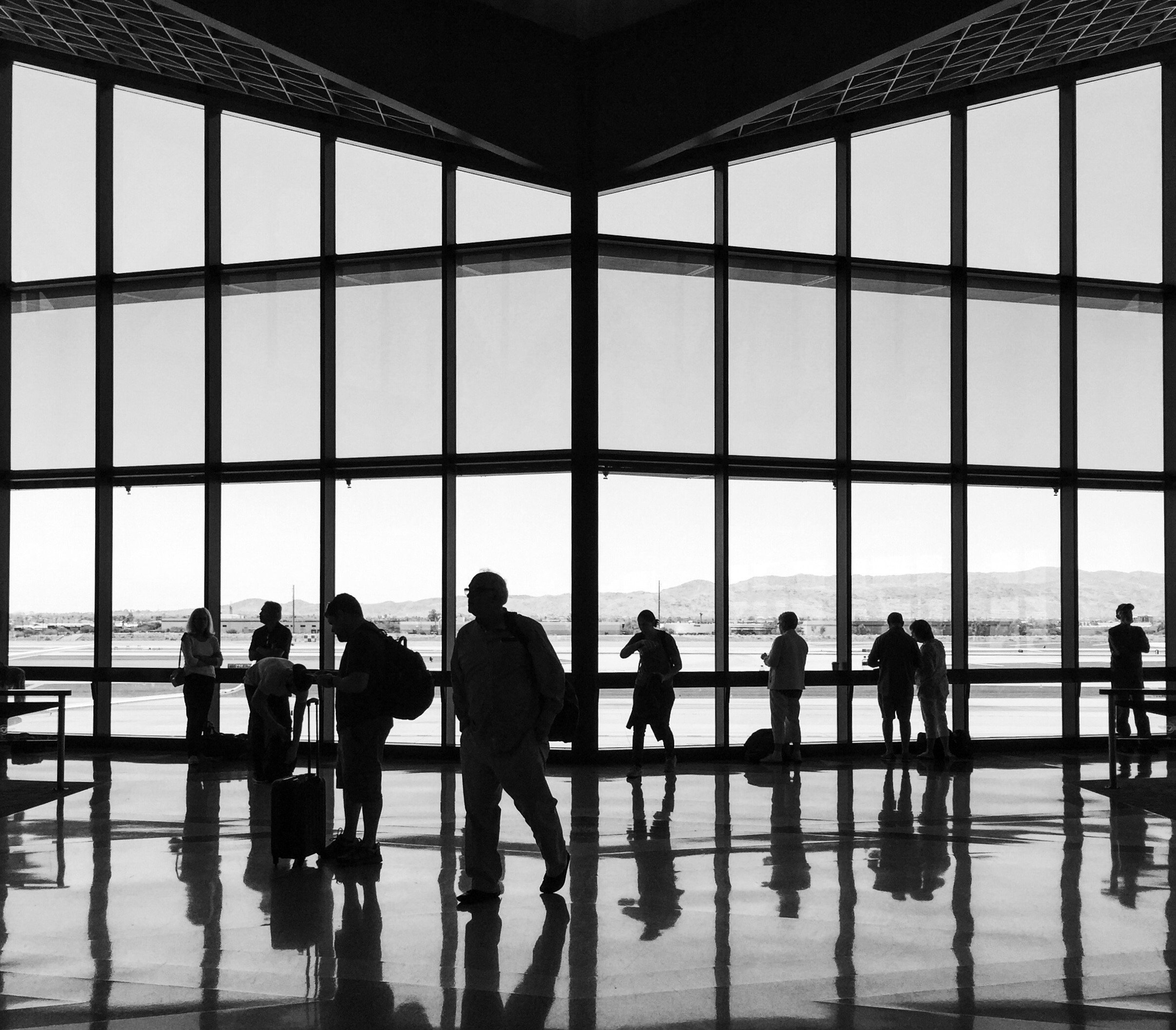 People waiting at an airport terminal with large glass windows showing the runway and mountains in the background, silhouetted against bright daylight.
