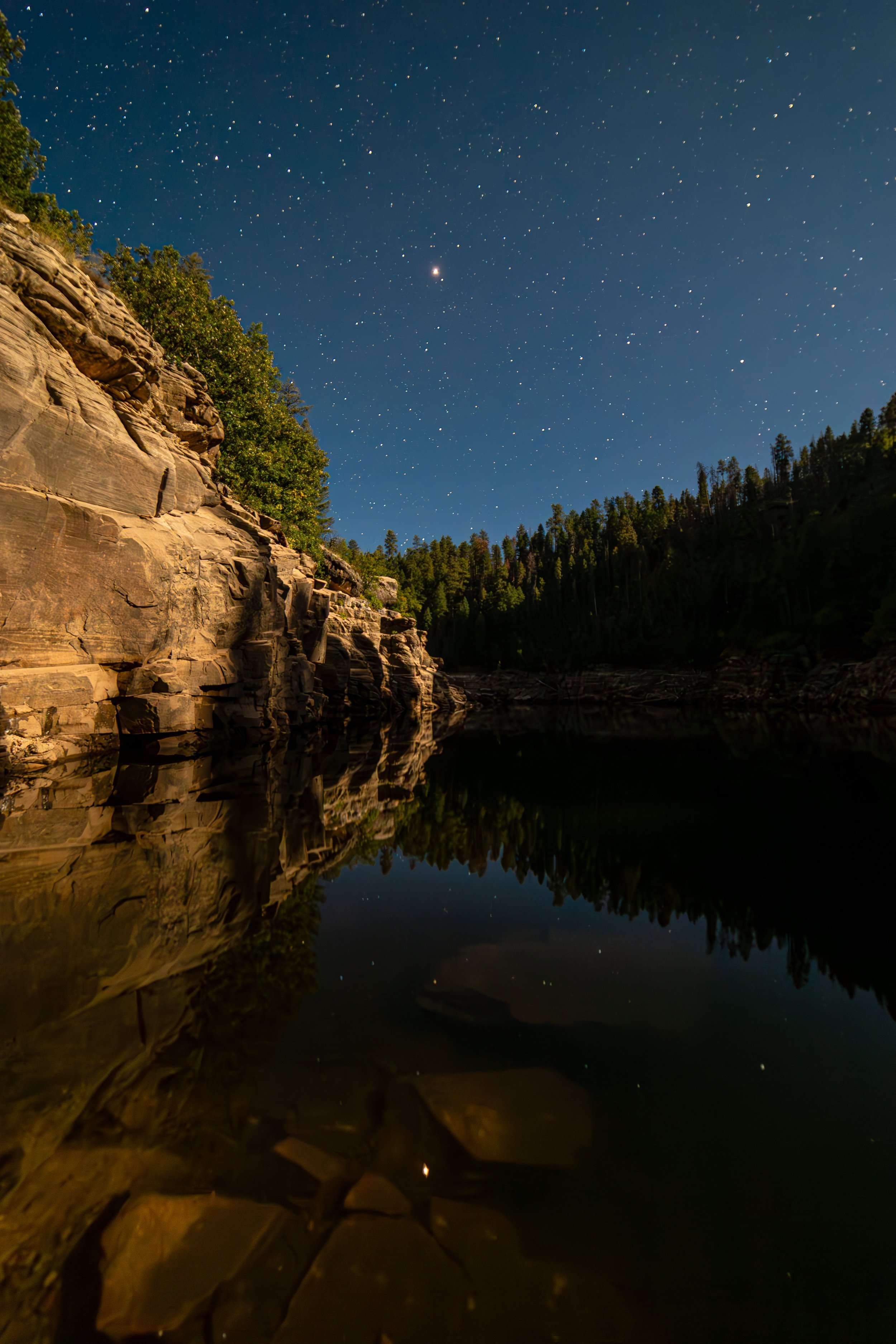 Nighttime landscape scene with a rocky cliff on the left, evergreen trees, and a clear starry sky reflected in a calm body of water.