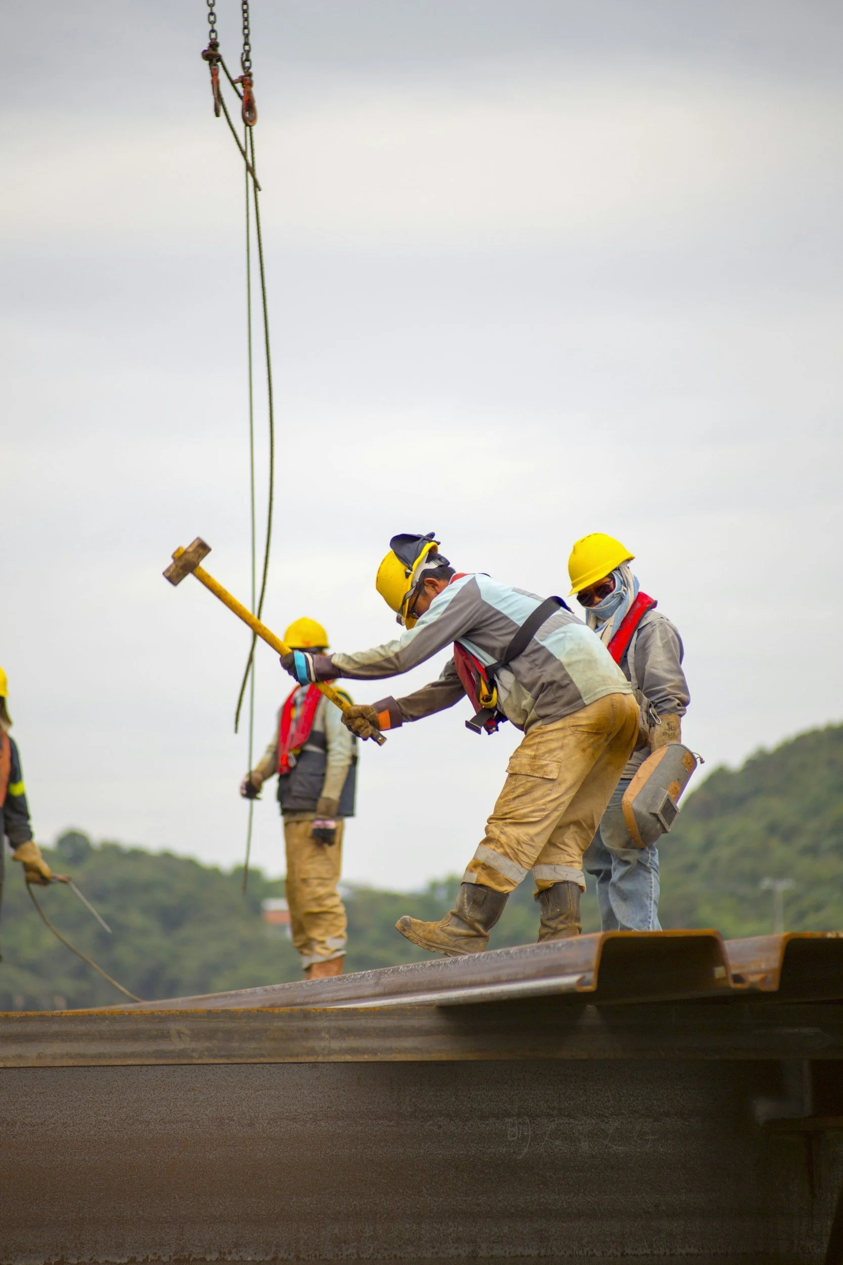 Construction workers wearing safety helmets and gear working on steel beam during construction, with a hilly landscape in the background.