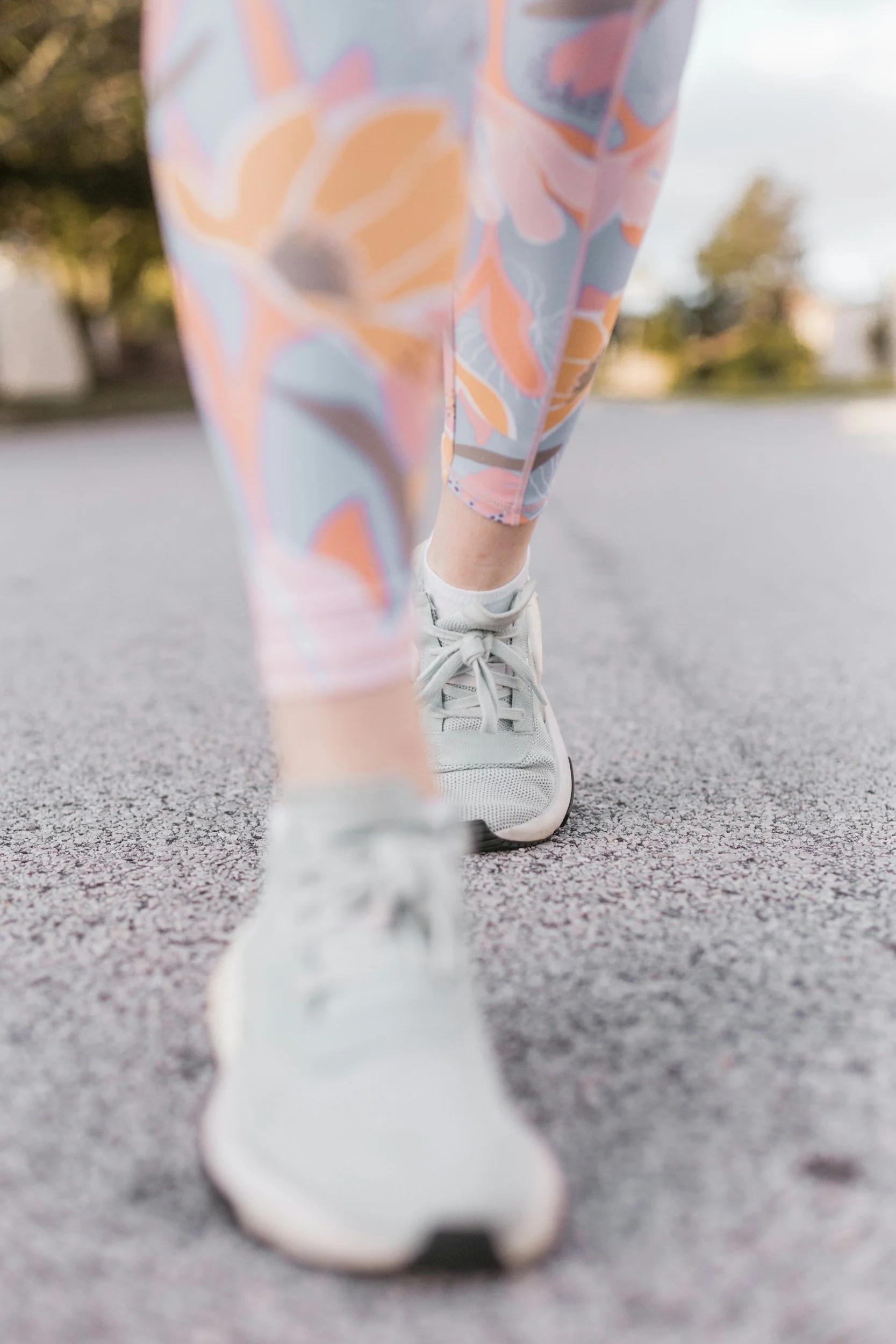Close-up of a person walking on pavement, wearing patterned leggings, white athletic shoes, and white socks, with a blurry background of trees and houses.