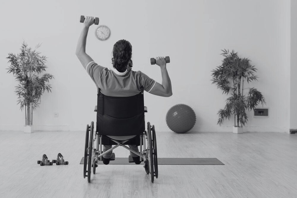 Person in a wheelchair lifting dumbbells in a fitness or therapy room, with plants and exercise equipment around.