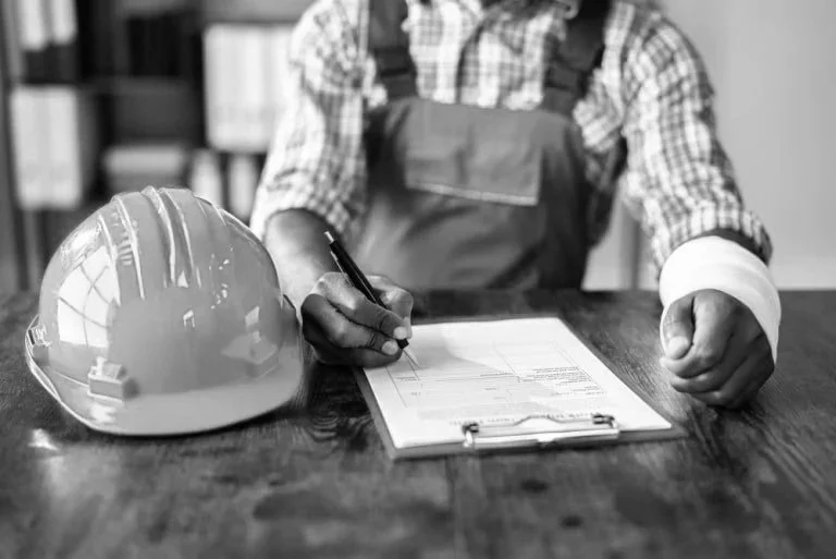 A person in work attire, possibly a construction worker, signs a document on a clipboard with a pen, with a safety helmet placed on the table.
