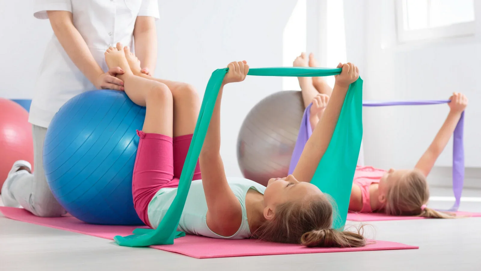 Two young girls using resistance bands while lying on yoga mats during a fitness class with an instructor assisting one girl with an exercise ball.