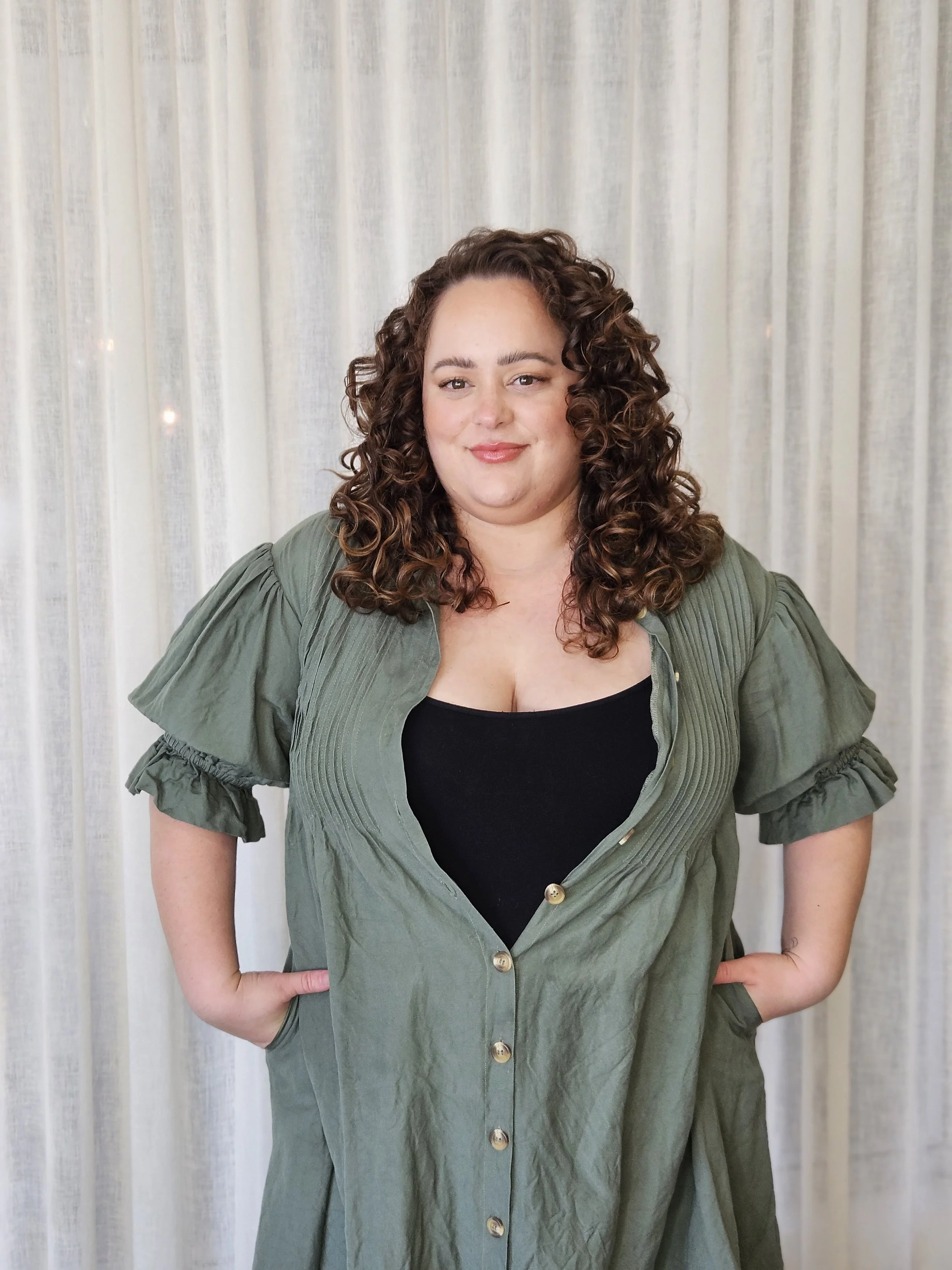 A woman with curly brown hair, wearing a black top and an olive-green button-up dress, standing in front of cream-colored curtains, smiling slightly with hands in her pockets.