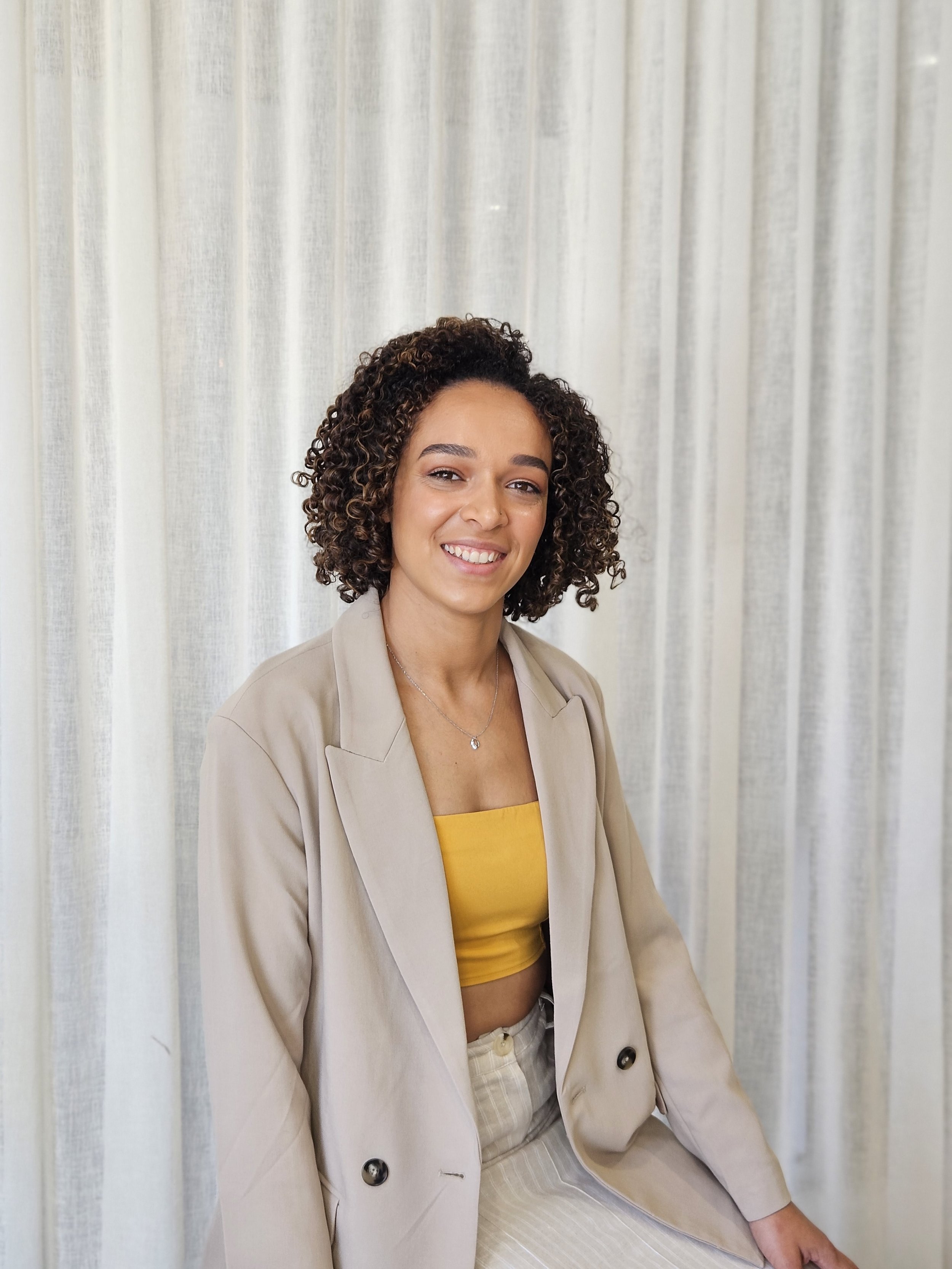 A young woman with curly hair, wearing a beige blazer over a yellow crop top, smiling at the camera with a light-colored curtain background.