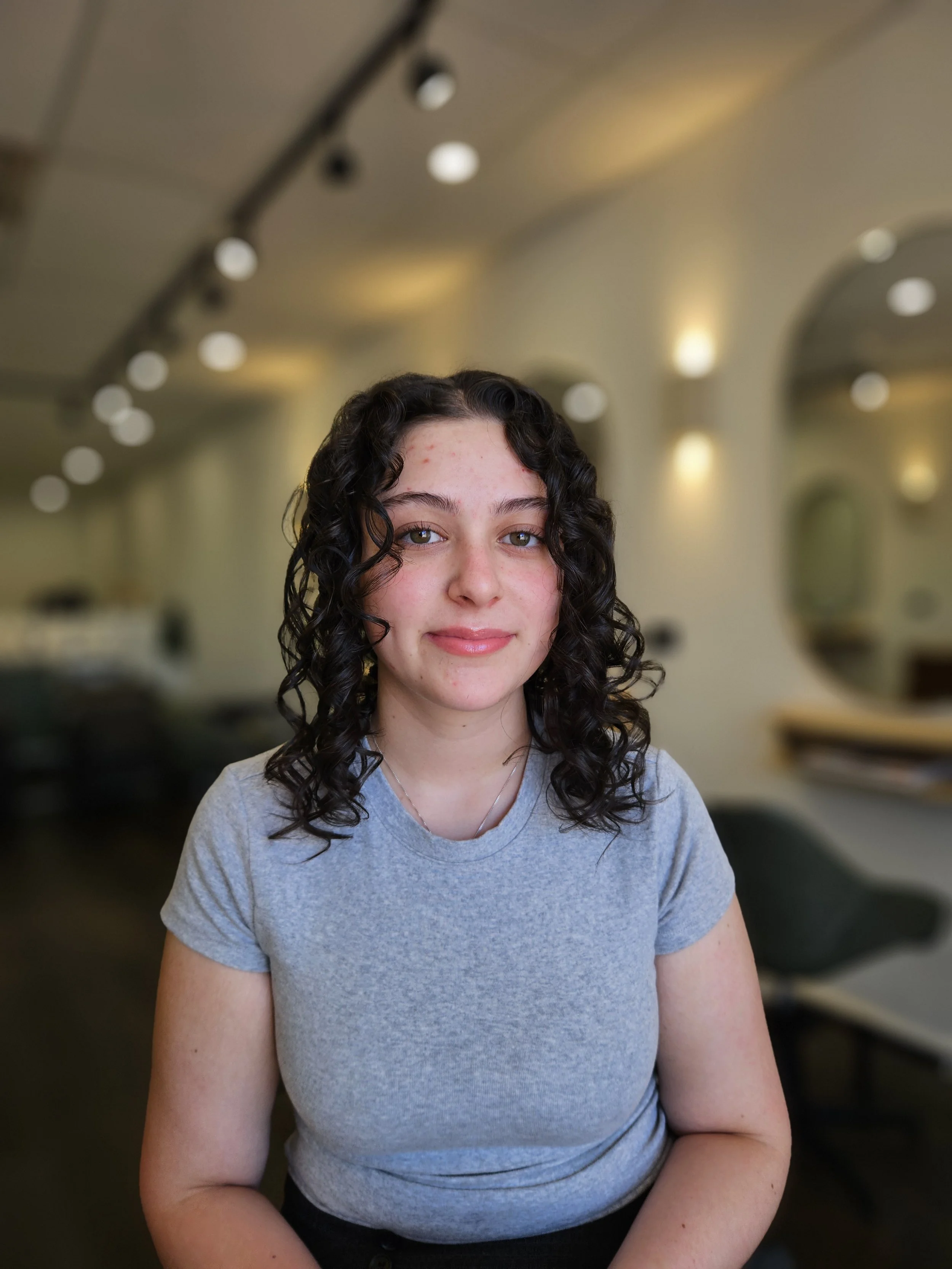 A young woman with curly dark hair and fair skin, wearing a gray t-shirt, sitting in a modern, softly lit room with blurred background.