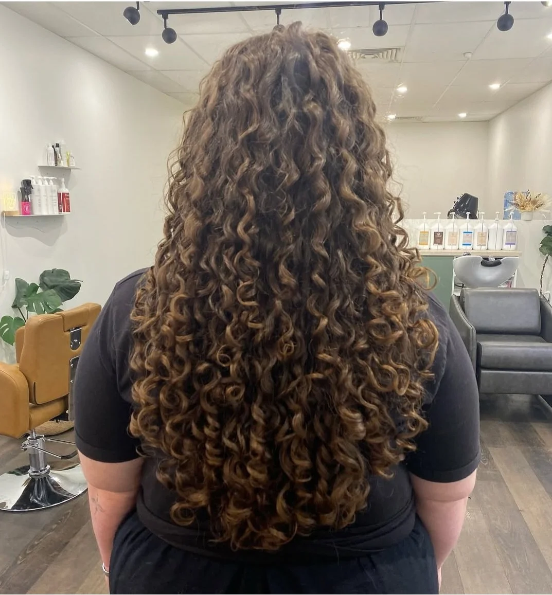 Back view of a woman with long, curly brown hair in a hair salon.