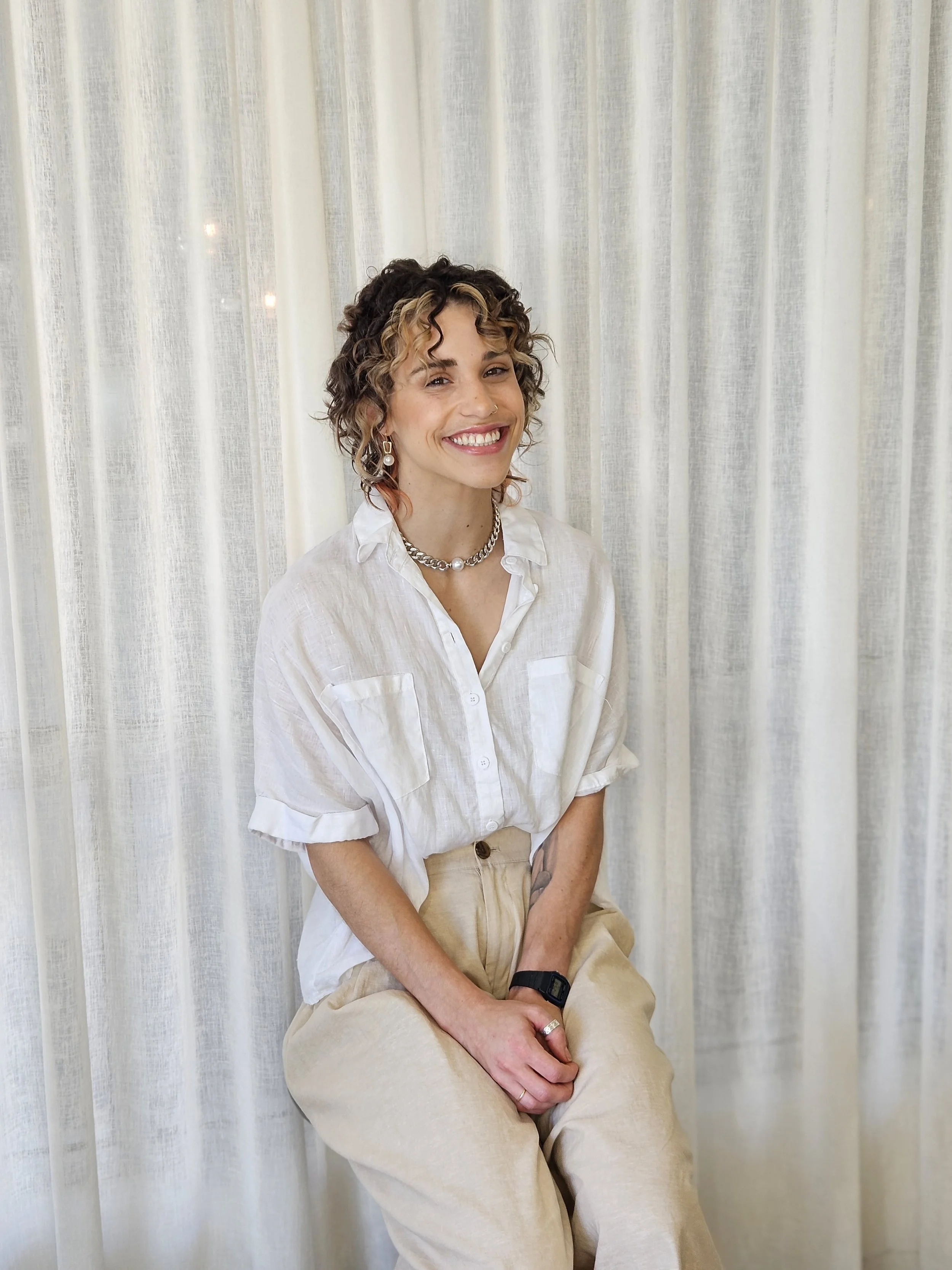 A woman with curly hair and a bright smile, wearing a white shirt and beige pants, sitting in front of a white curtain.