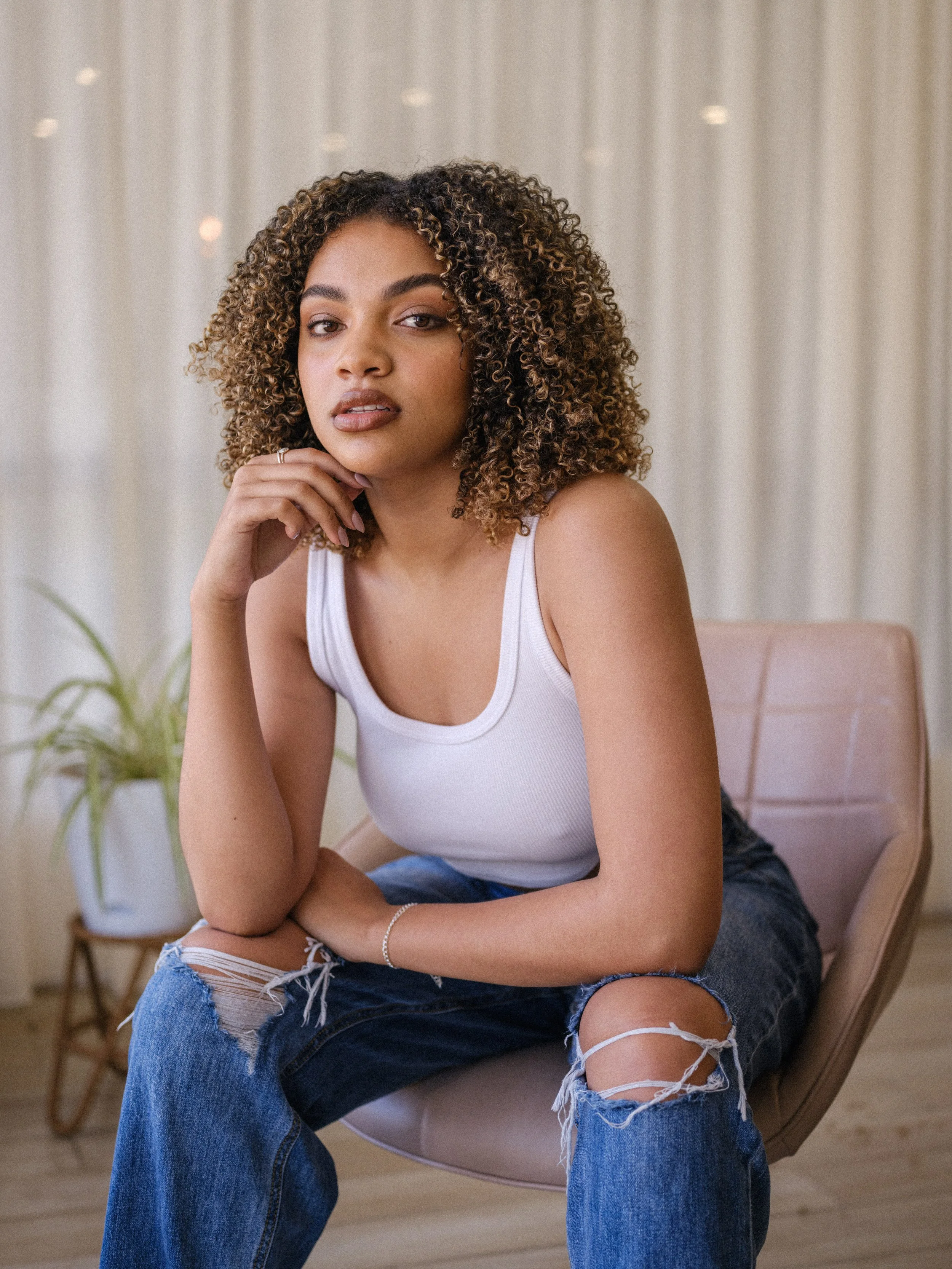 A young woman with curly hair wearing a white tank top and ripped jeans, sitting in a modern room with light-colored curtains and a potted plant in the background.