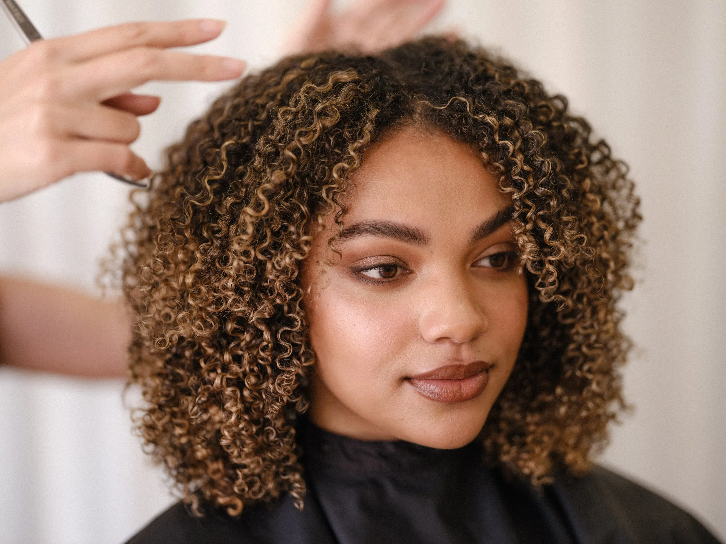 A woman with short, curly hair in shades of brown, receiving a styling treatment from a stylist with a hand near her hair, in a salon setting.