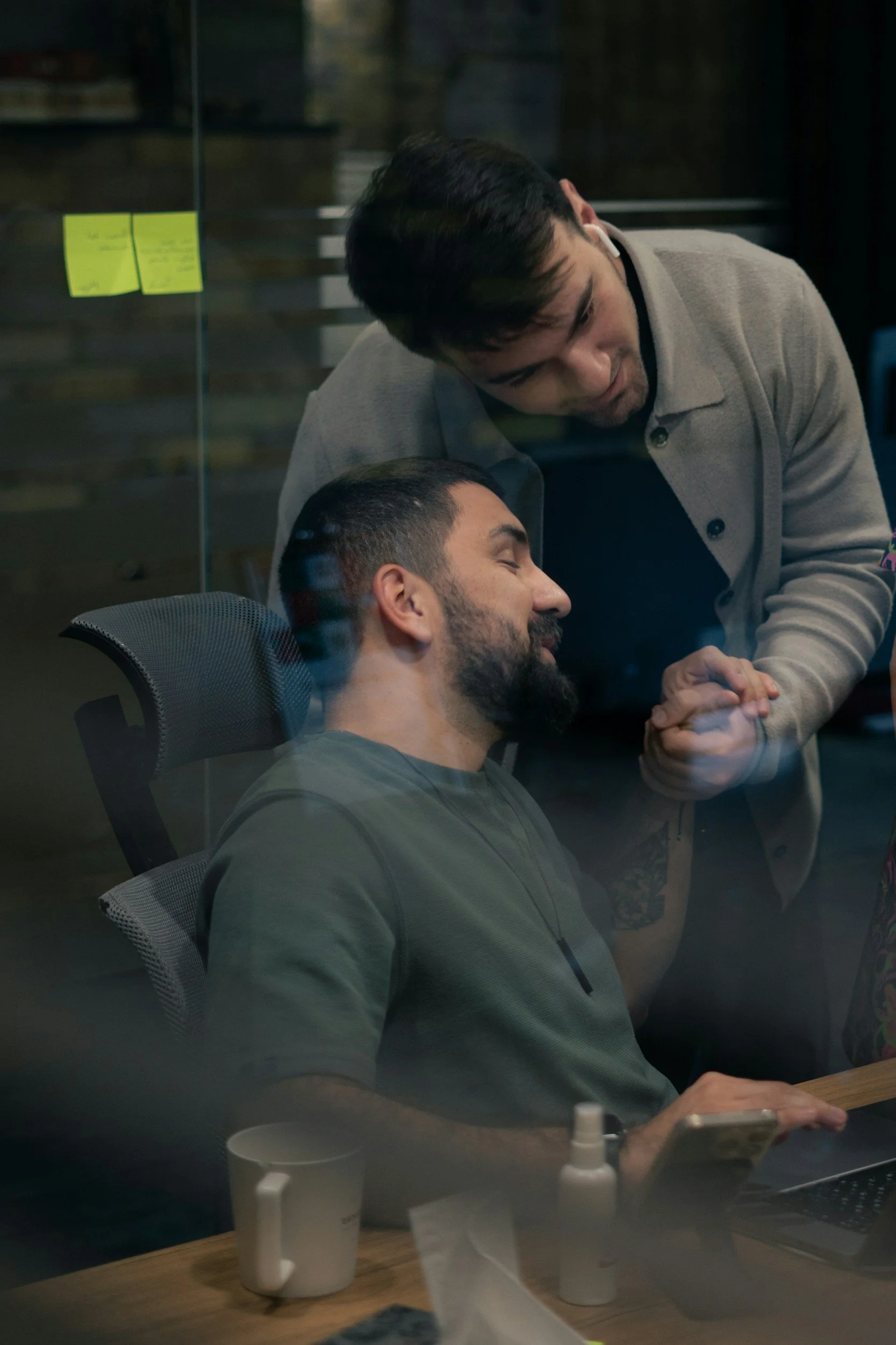 Two men interacting through a glass window. One is sitting at a desk with a coffee mug, phone, and laptop, while the other is standing and holding his hand. They appear to be sharing a moment of connection.