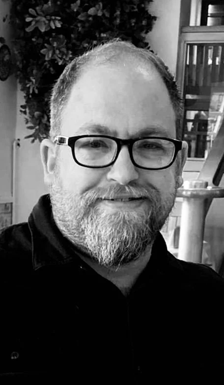 Black and white photo of a smiling man with glasses and a beard, sitting indoors near a plant and a staircase.