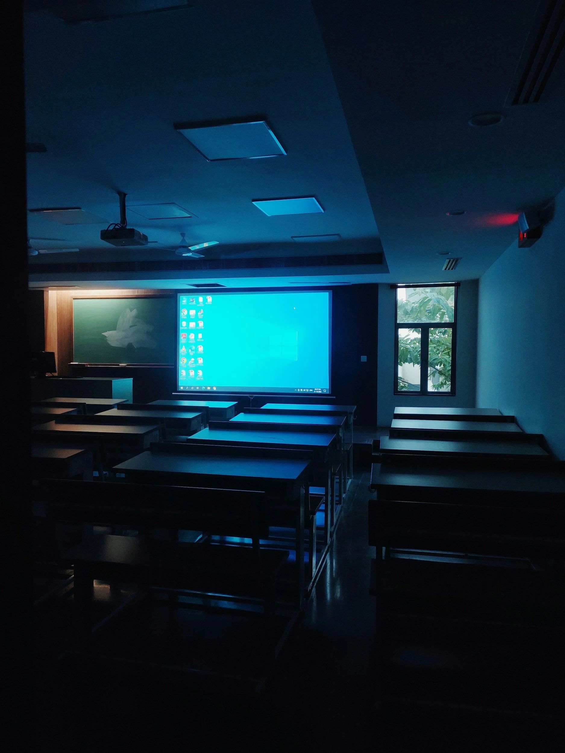 Empty classroom with chairs and desks, illuminated by blue light from a projector screen displaying a Windows desktop, a chalkboard, and a window showing outside greenery.