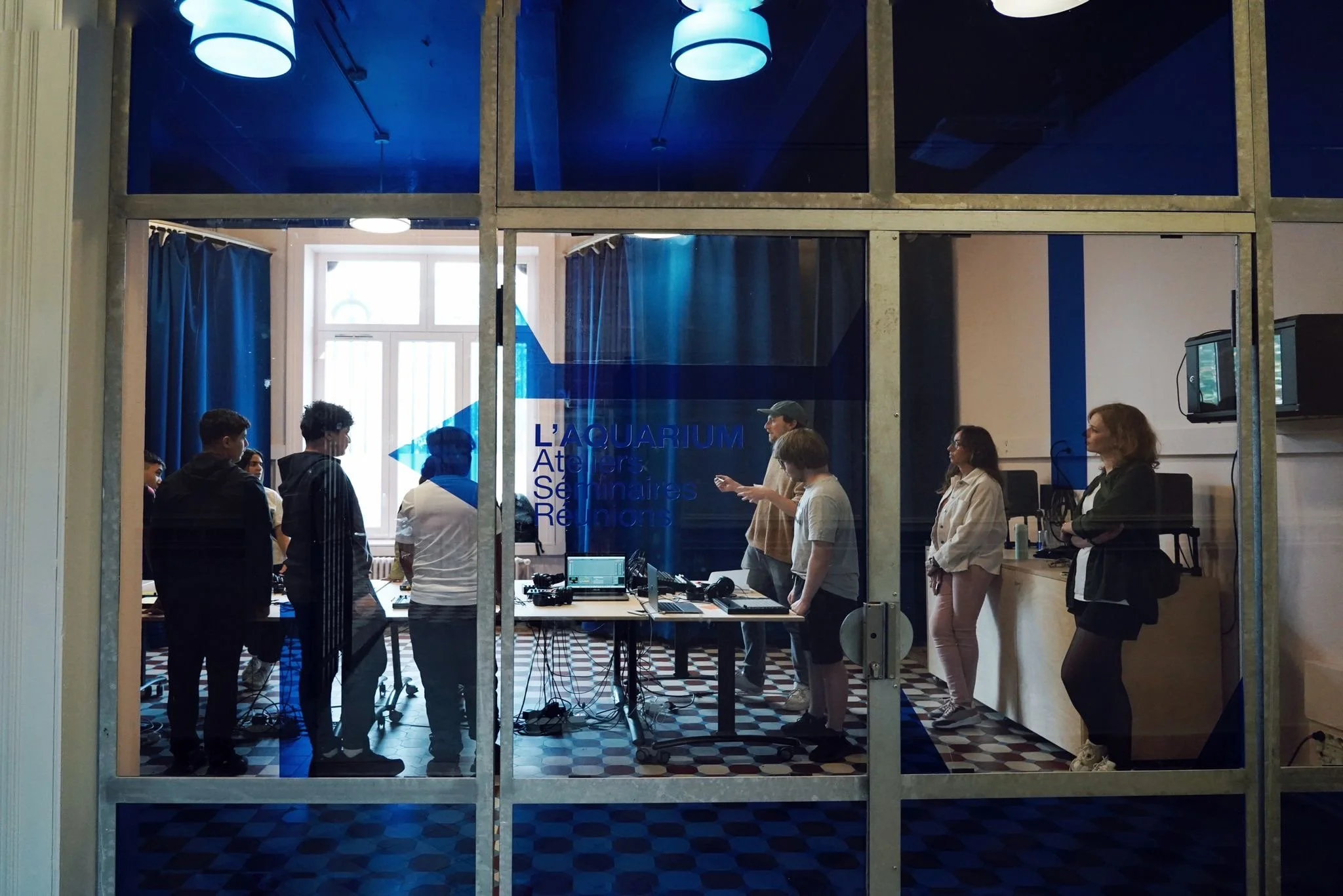 A group of people inside a room with computers and audio equipment, viewed through a window with metal framing and blue-tinted glass. The room has a high window, blue curtains, and a sign that reads 'L'AQUARIUM Atelier Seines Réseaux' on the wall.