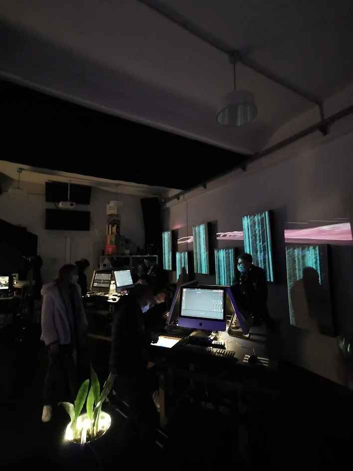 A dimly lit room with multiple people operating computers and digital screens, and abstract digital graphics projected on the walls.