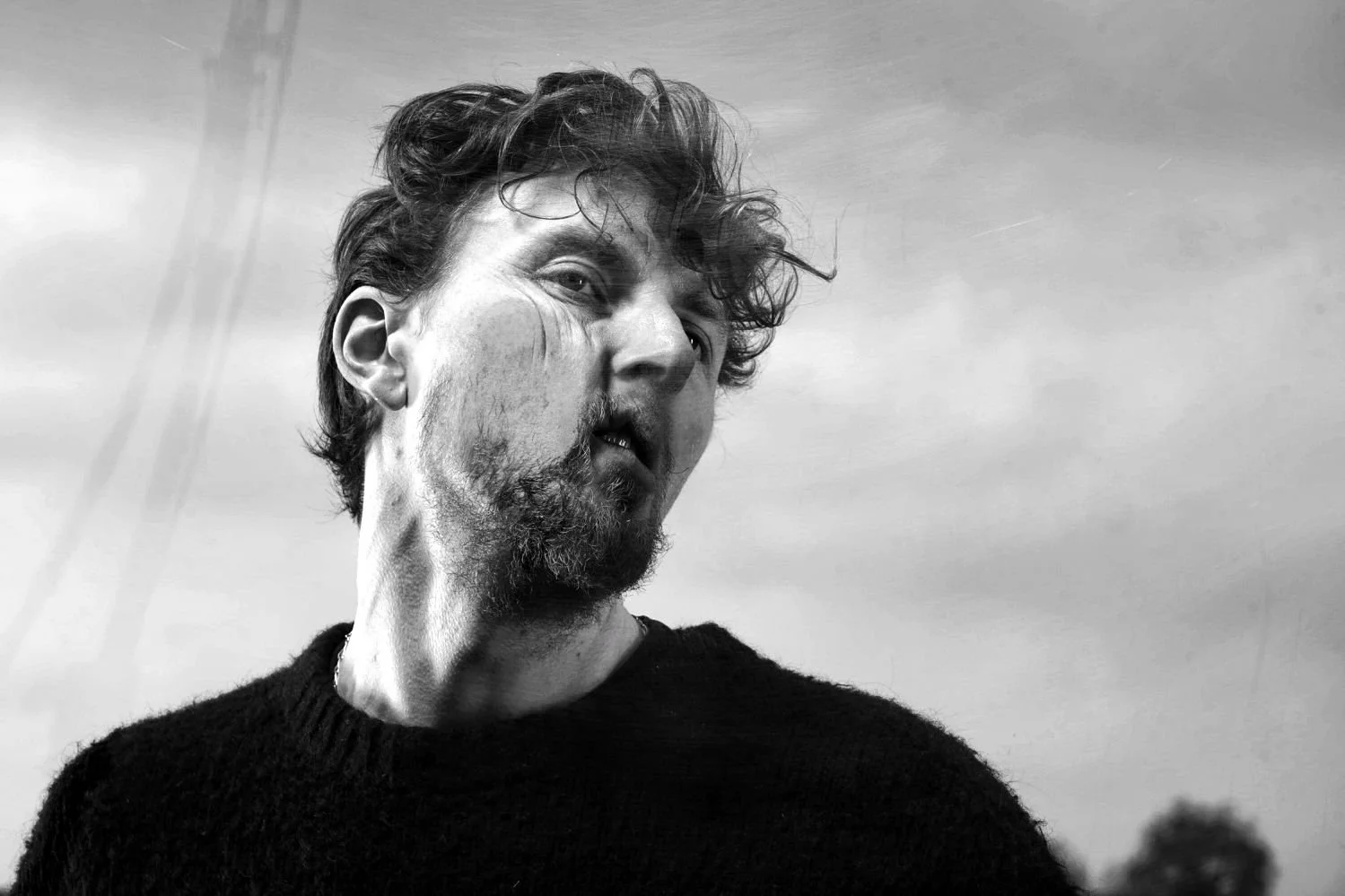 Black-and-white photo of a man with curly hair, a beard, and a mustache, looking to his left with a slightly open mouth, wearing a dark sweater, outdoors against a cloudy sky background.