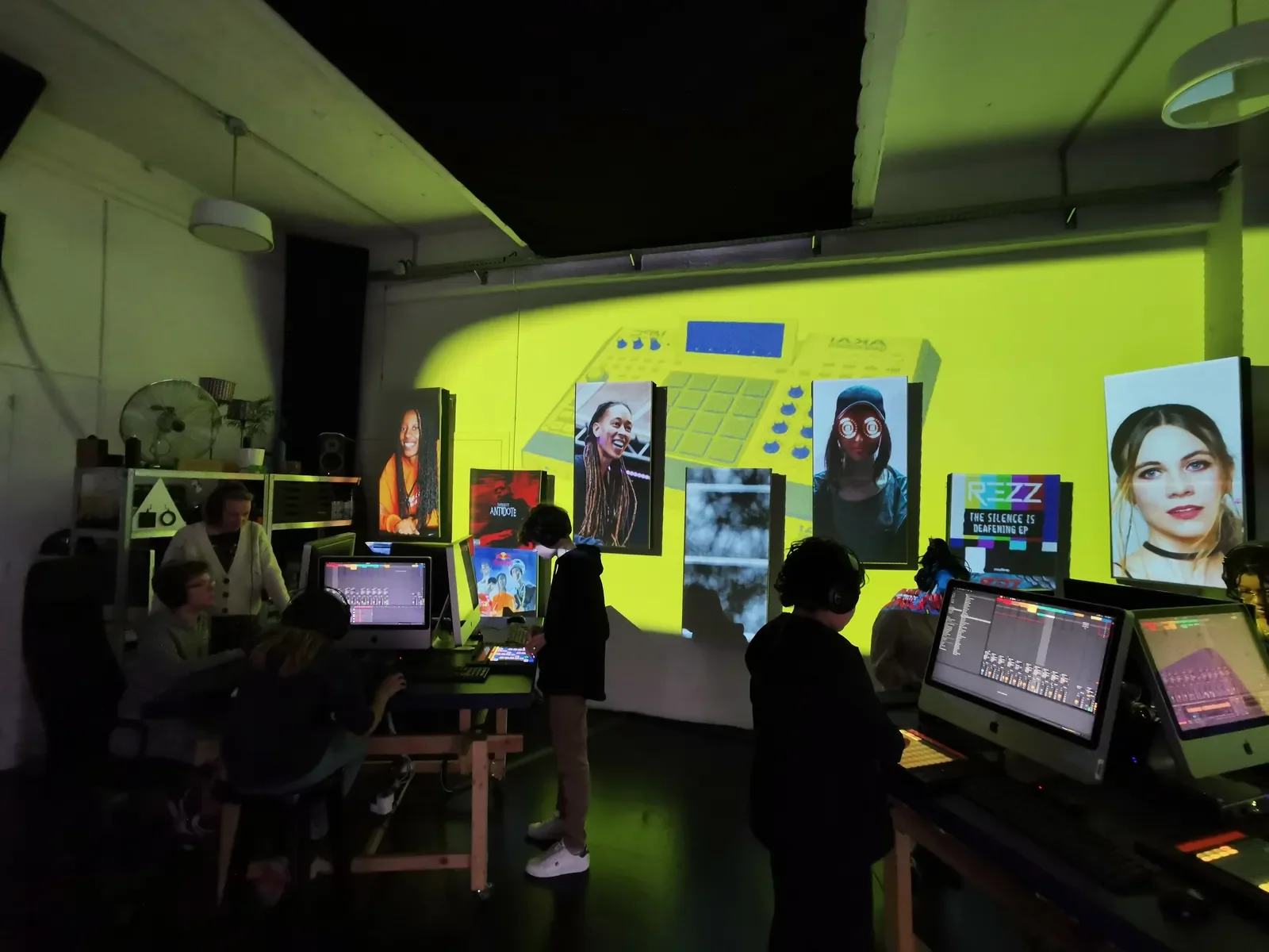 A group of people working in a music or media production studio with large screens displaying images and graphics, including portraits of women, and a yellow background with a graphic of a music production device.