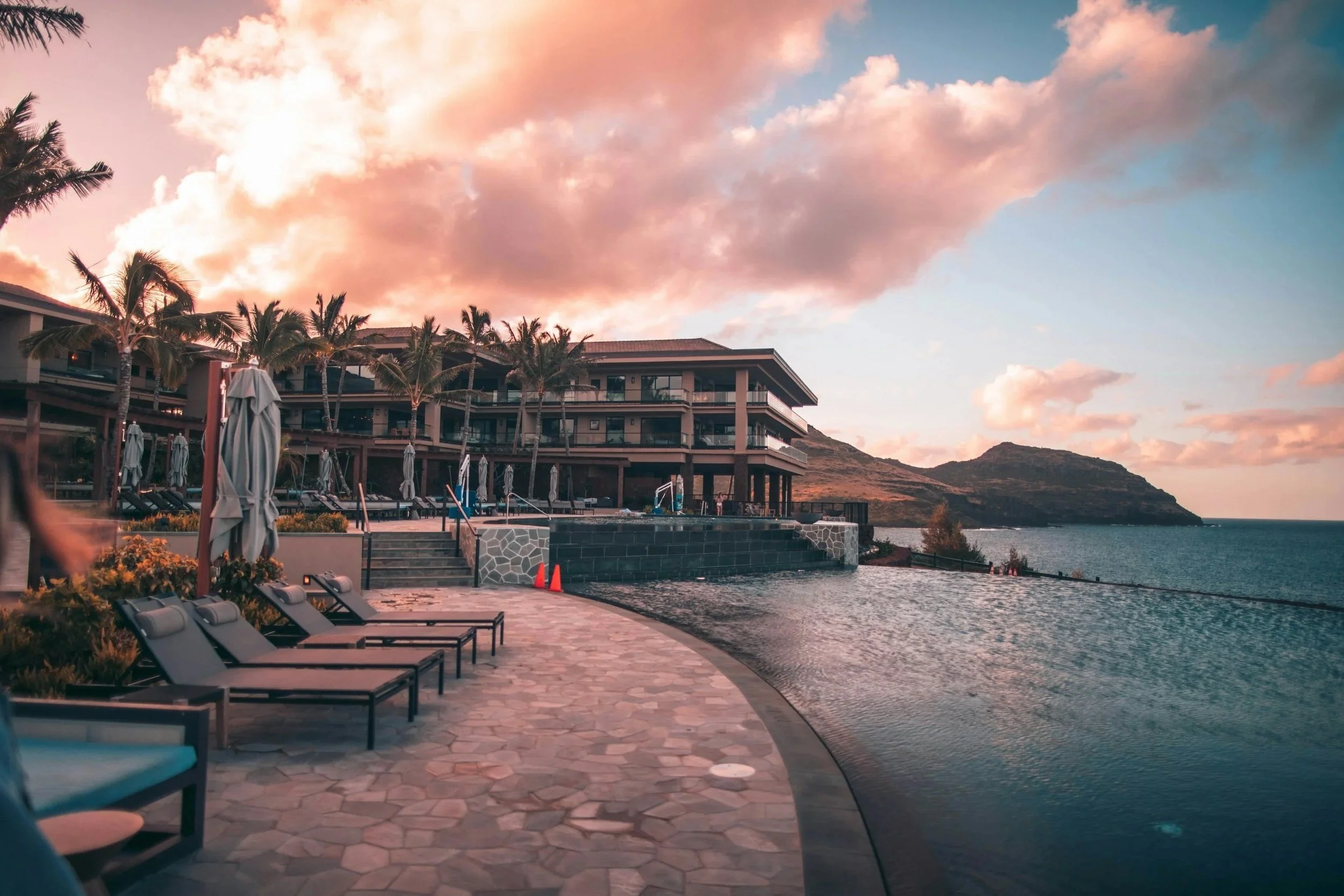 A luxurious seaside resort at sunset with an infinity pool, lounge chairs, and a building with multiple balconies, overlooking the ocean and mountains in the background.