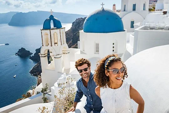 Smiling couple in sunglasses on white terrace with blue-domed churches overlooking the sea in Santorini, Greece.