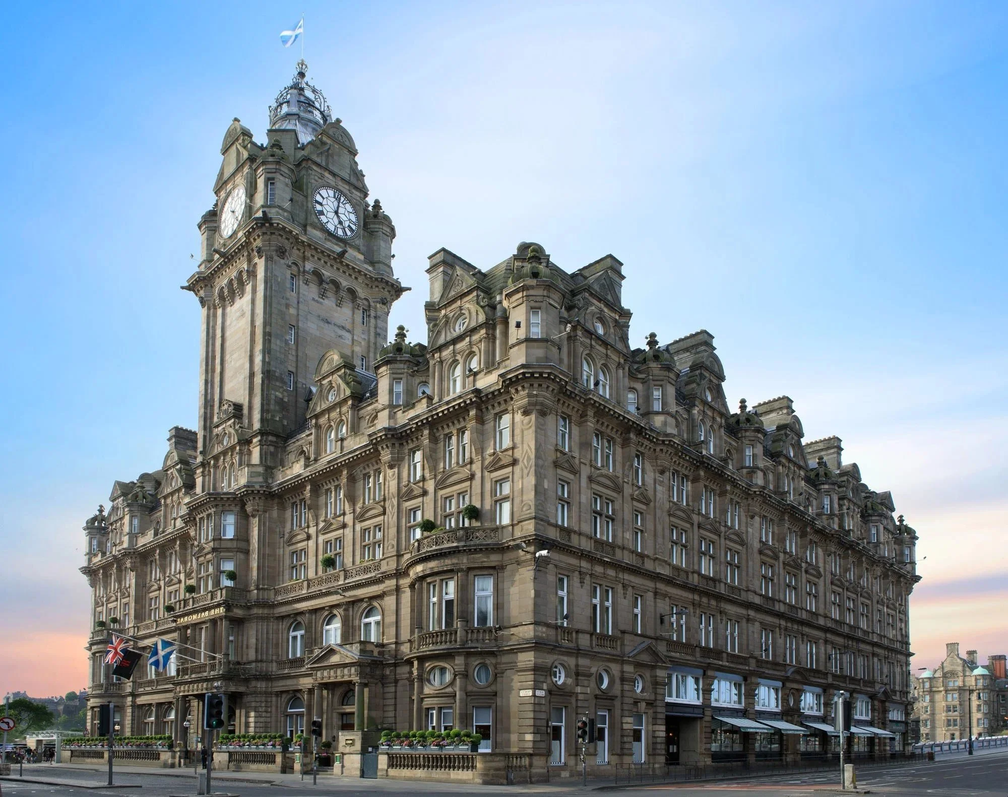 Historic multi-story building with a clock tower, flags, street lamps, and a corner entrance, set against a blue sky with some clouds.