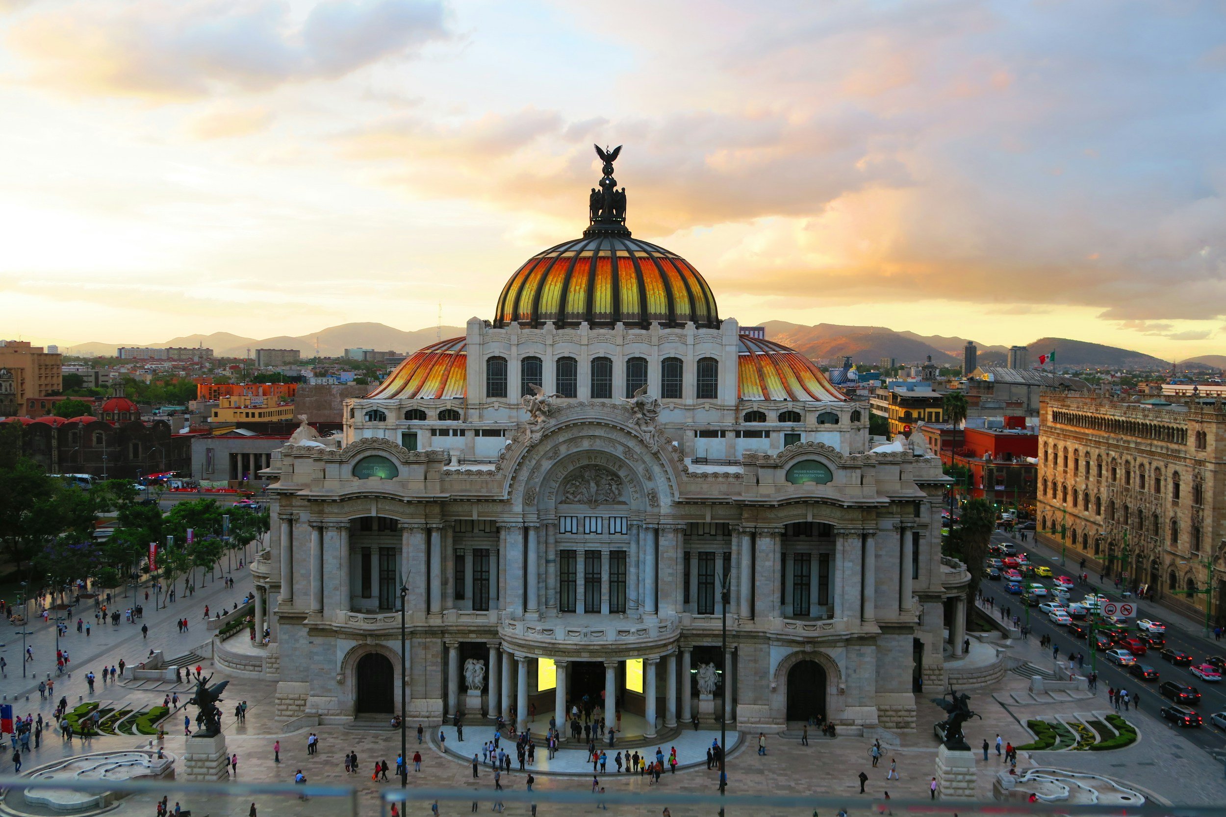 Exterior view of the Palace of Fine Arts in Mexico City during sunset with a golden dome and people walking in front.